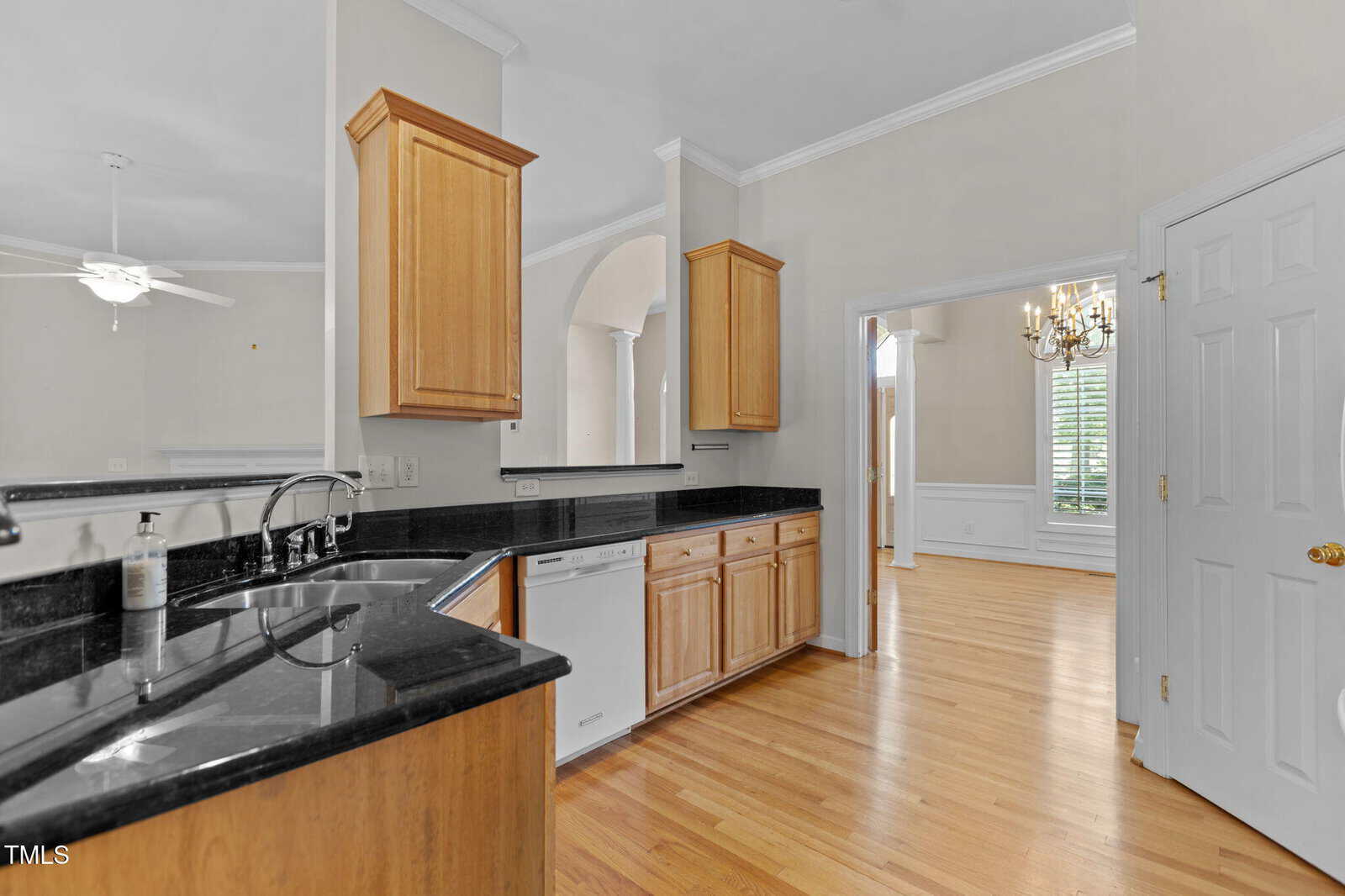 A kitchen with light wood cabinets, black granite countertops, a stainless steel sink, dishwasher, and light-colored hardwood floors. The kitchen opens to a dining area with a chandelier and large window.