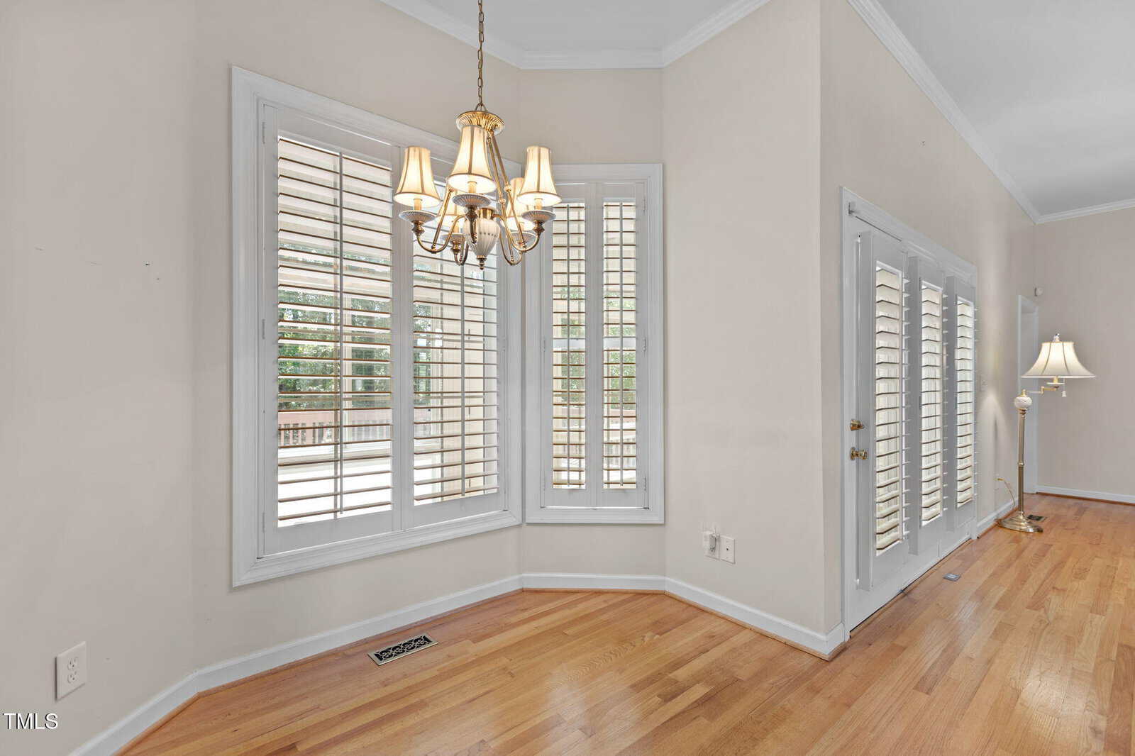 Bright room with large bay windows, white plantation shutters, a brass chandelier, hardwood floors, and double glass doors leading outside. Walls are light beige, and there is a floor lamp in the background.