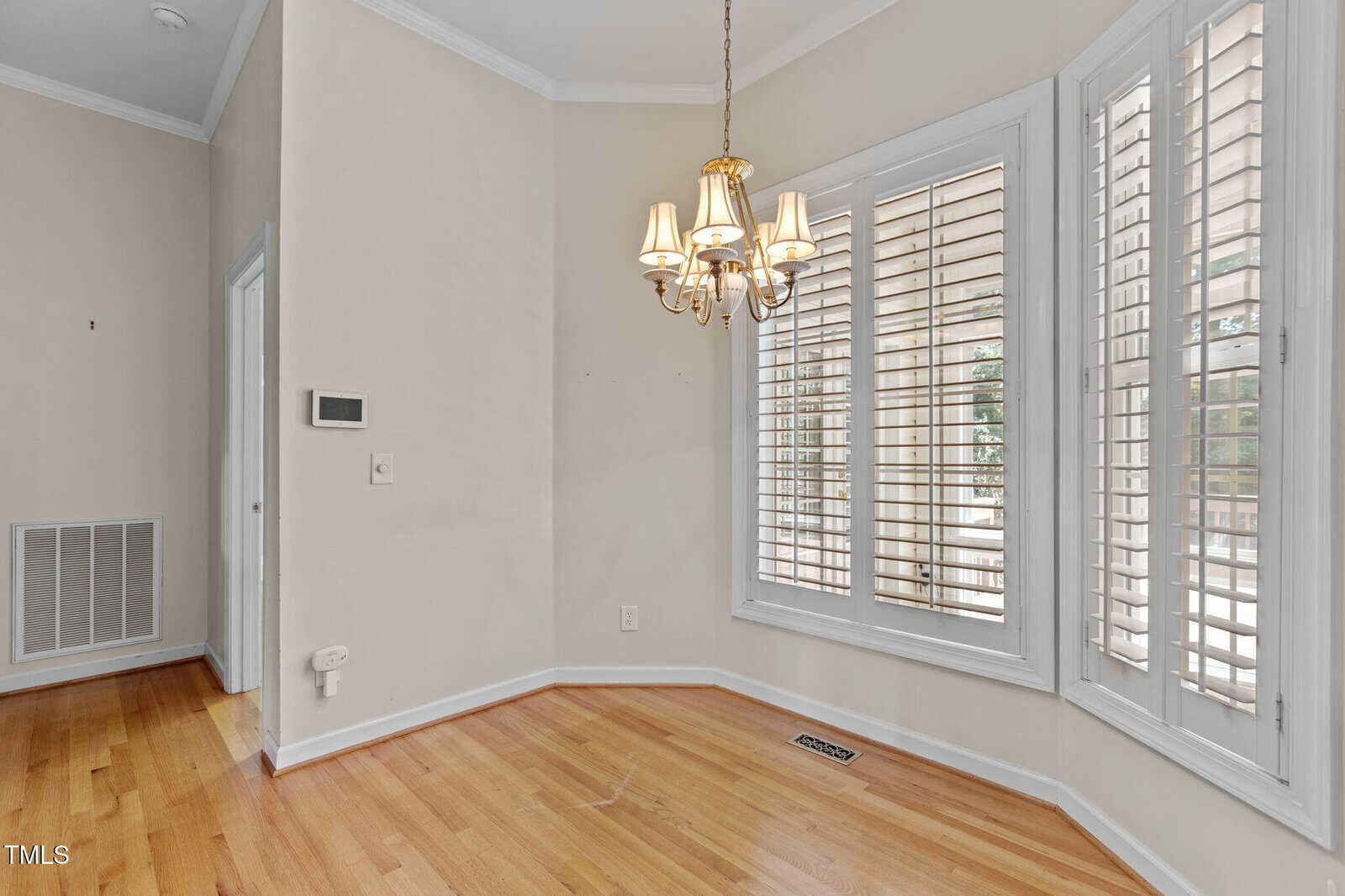 A corner room with light hardwood floors, beige walls, a brass chandelier, and large windows with white plantation shutters letting in natural light.