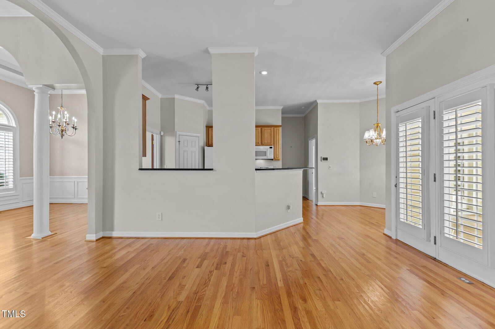 Open kitchen and dining area with hardwood floors, white walls, crown molding, arched doorway, windows with shutters, and French doors leading outside. Chandeliers hang in both dining spaces.