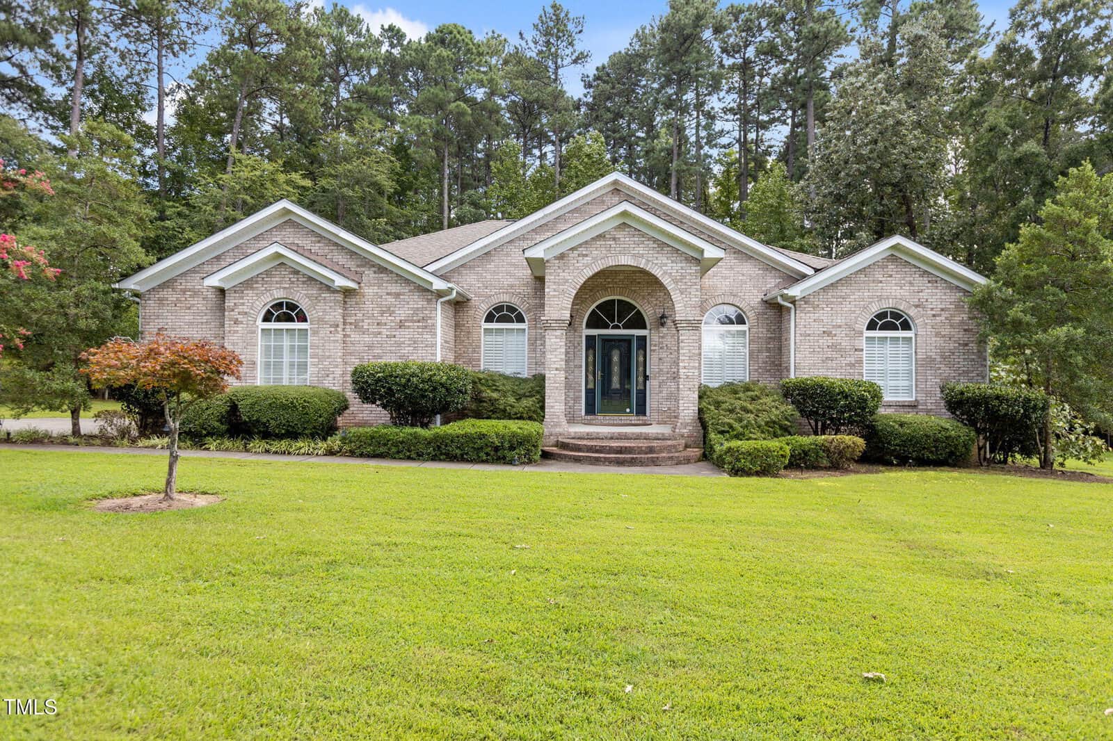 Charming single-story brick house at 139 Ridge Road in Butner, featuring arched windows, white trim, and a covered entry with columns. Enjoy a manicured lawn, shrubs, and tall trees providing a picturesque backdrop.