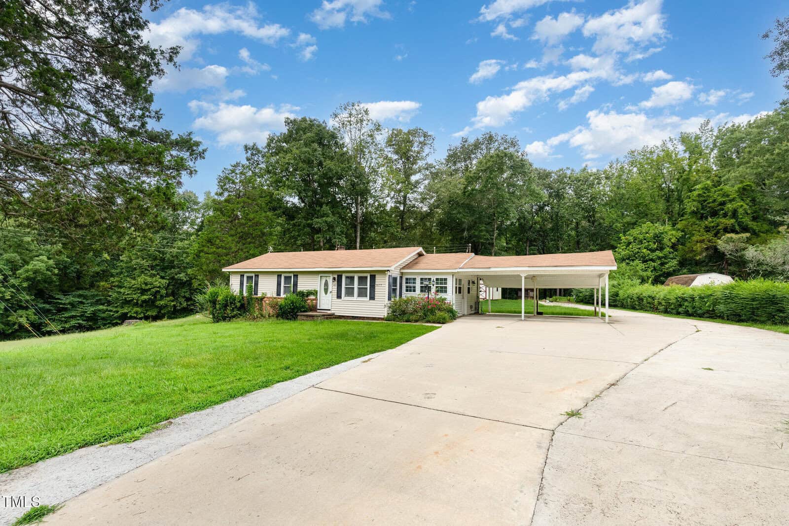 Single-story house with tan siding and a brown roof, featuring a covered carport and a wide concrete driveway. The yard is grassy, and the property is surrounded by trees under a partly cloudy sky.