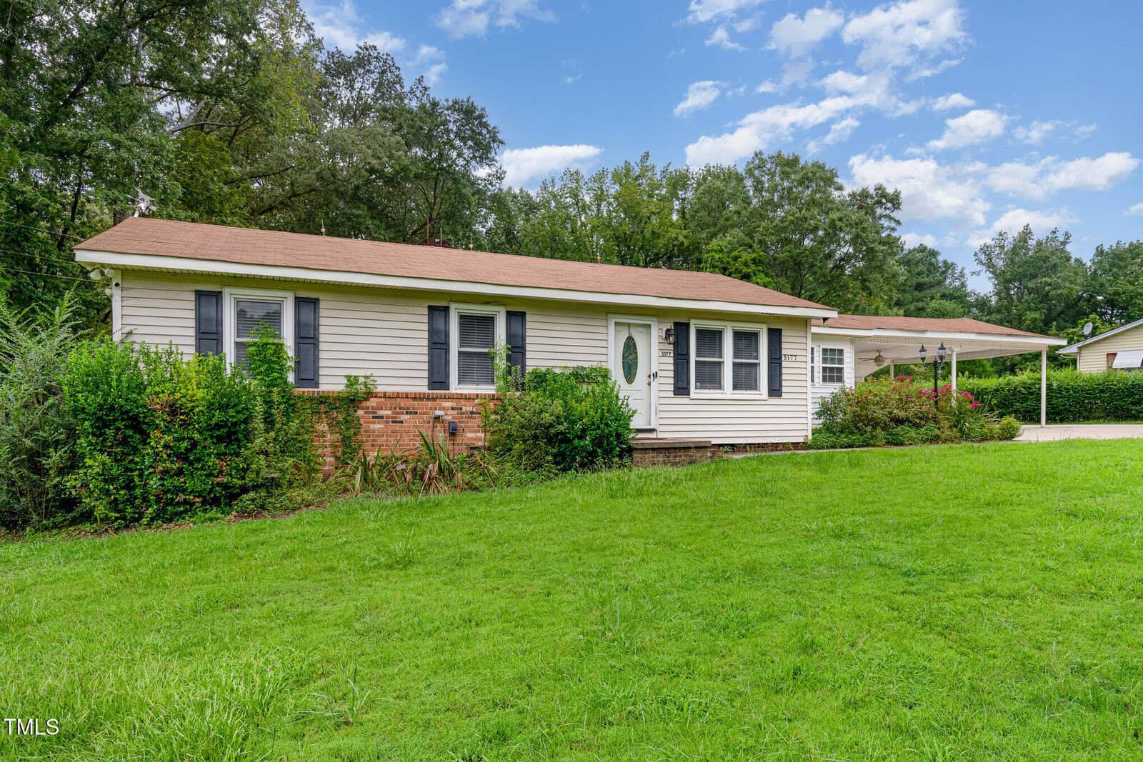 Single-story house with beige siding, brick accents, and dark shutters. There is a covered carport on the right side, green grass lawn in front, and trees in the background under a partly cloudy sky.
