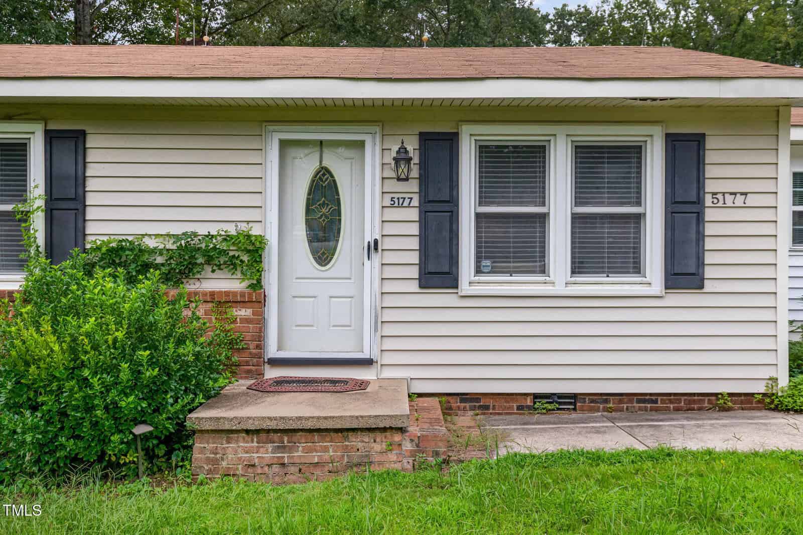 The image shows the front entrance of a beige house with black shutters, a white door with an oval window, brick steps, a concrete walkway, and green shrubs to the left of the door. The house number 5177 is visible.
