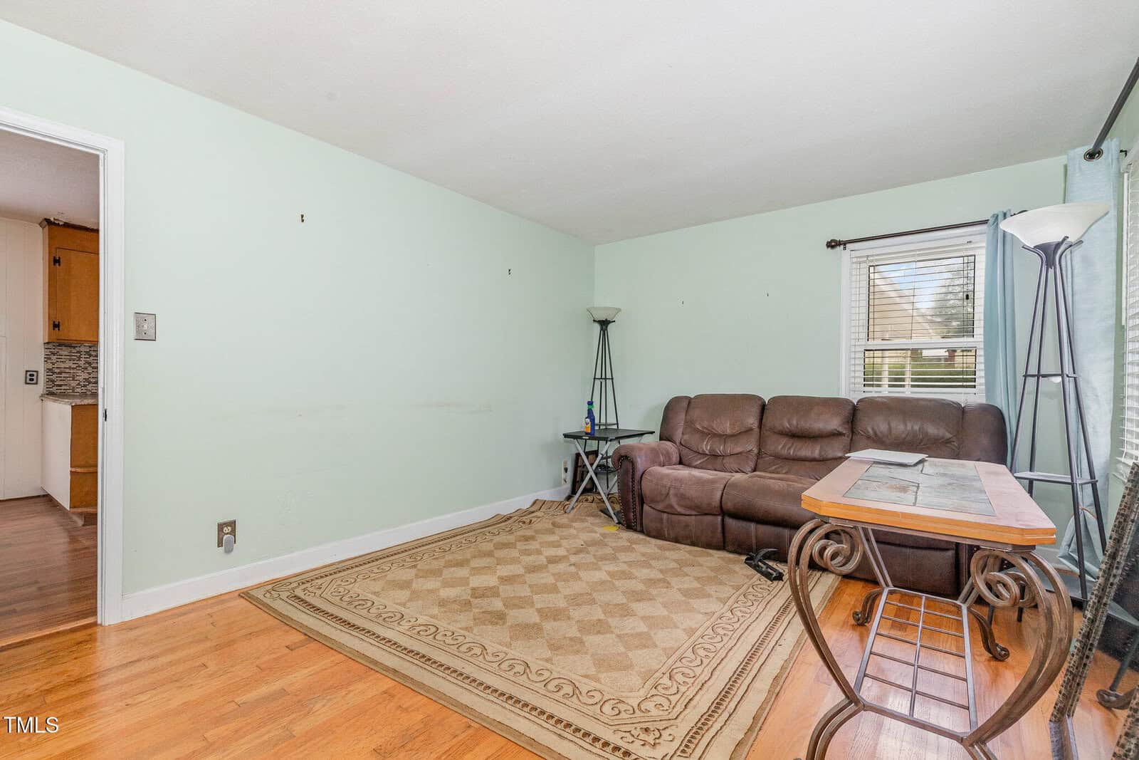 A living room with light green walls, a brown leather sofa, a patterned beige rug, two floor lamps, a glass-top table, and hardwood floors. A window with blinds lets in natural light.
