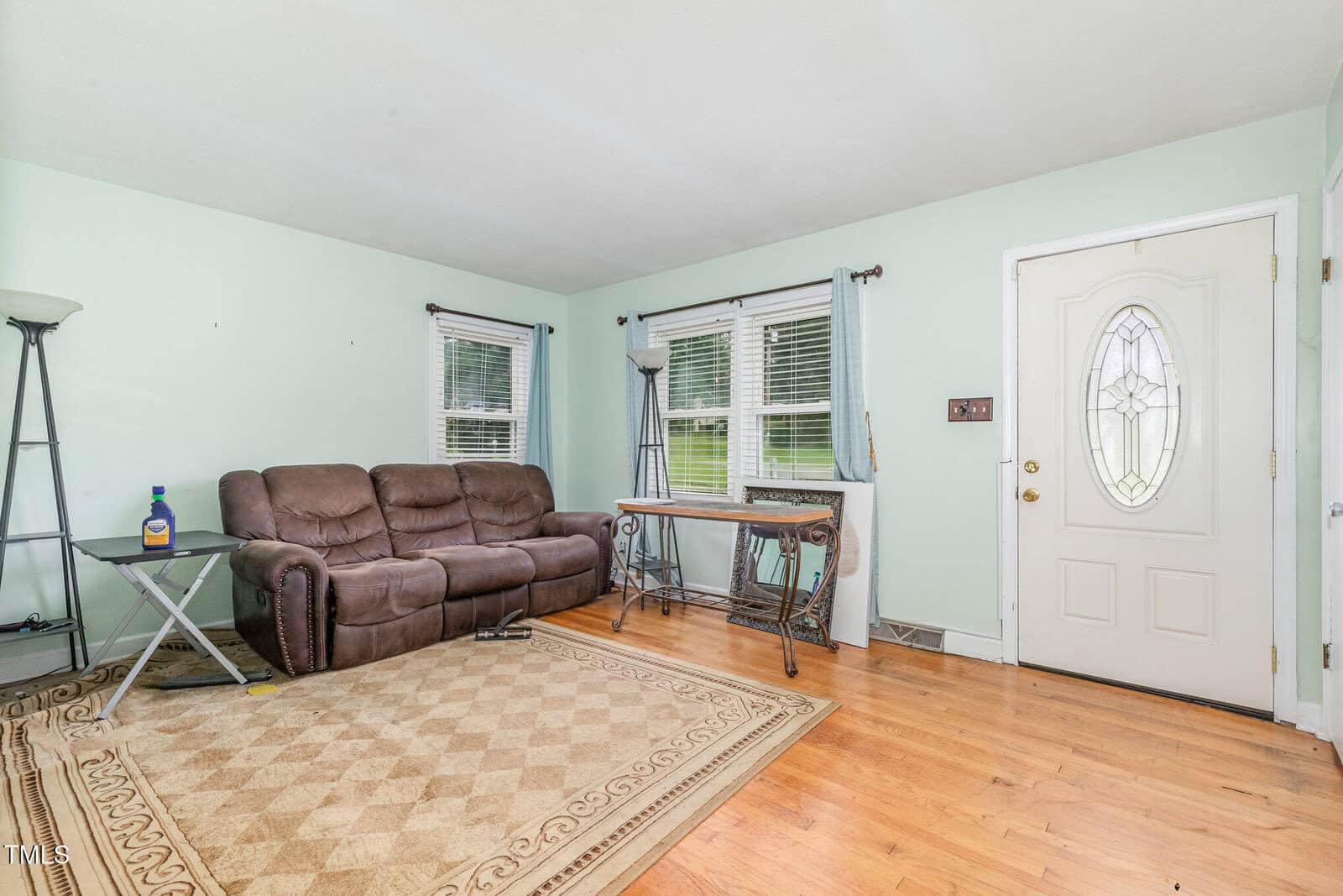 A living room with light green walls, wooden floors, a beige patterned rug, a brown reclining sofa, two floor lamps, a small table with a bottle on it, a console table, and a white front door with a glass panel.