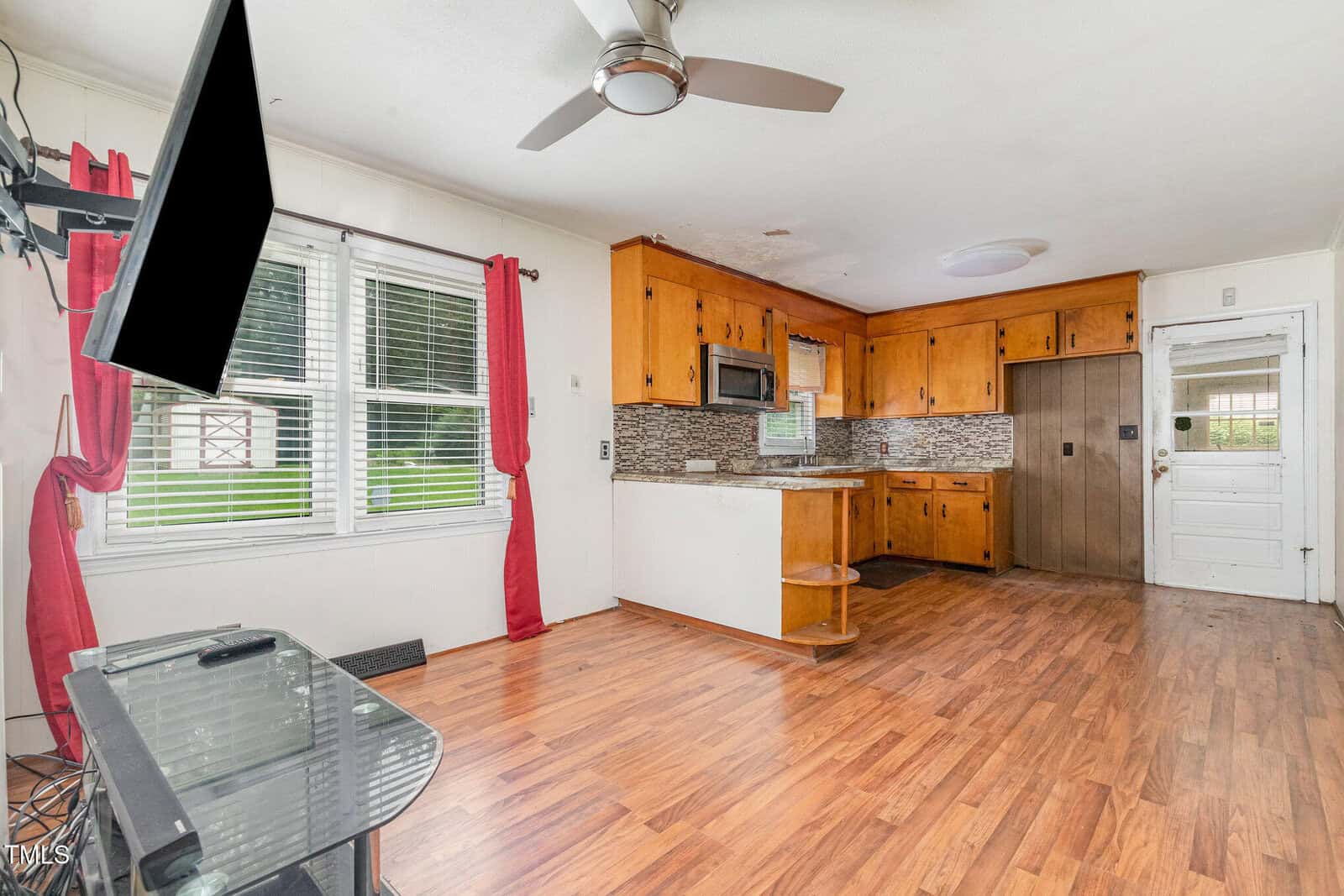 Open kitchen and living area with wood floors, wooden cabinets, a mounted TV, ceiling fan, tiled backsplash, window with red curtains, white walls, and a white back door with glass panels.