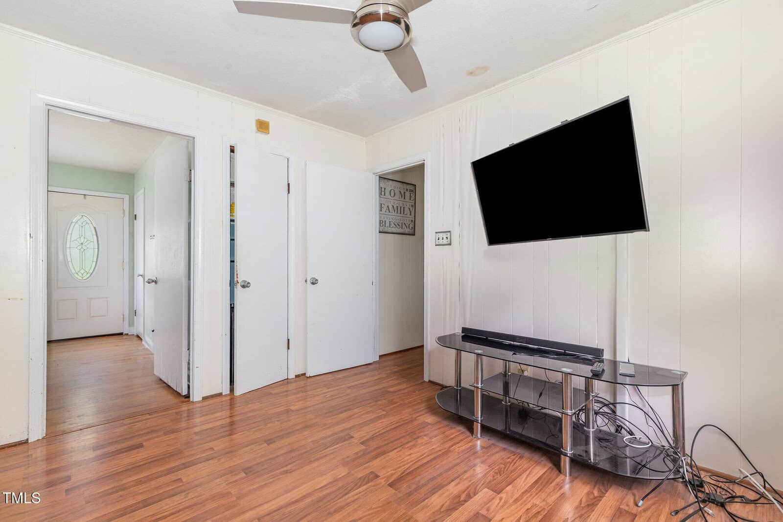 A room with hardwood floors, three white doors, a ceiling fan, a flat-screen TV mounted on the wall, a glass TV stand, and visible cables on the floor. A hallway with a decorative door is in the background.