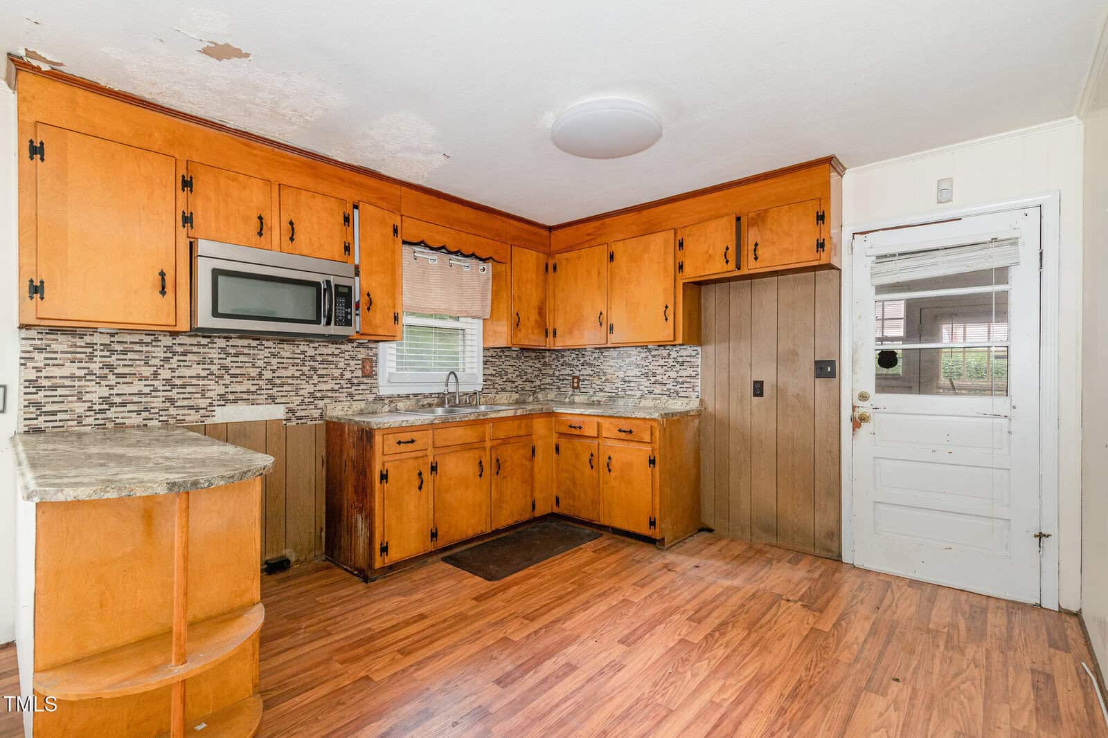 A kitchen with wood cabinets, a microwave, a window above the sink, laminate flooring, a mosaic tile backsplash, and visible ceiling and wall damage. There is a white door with glass panels on the right.