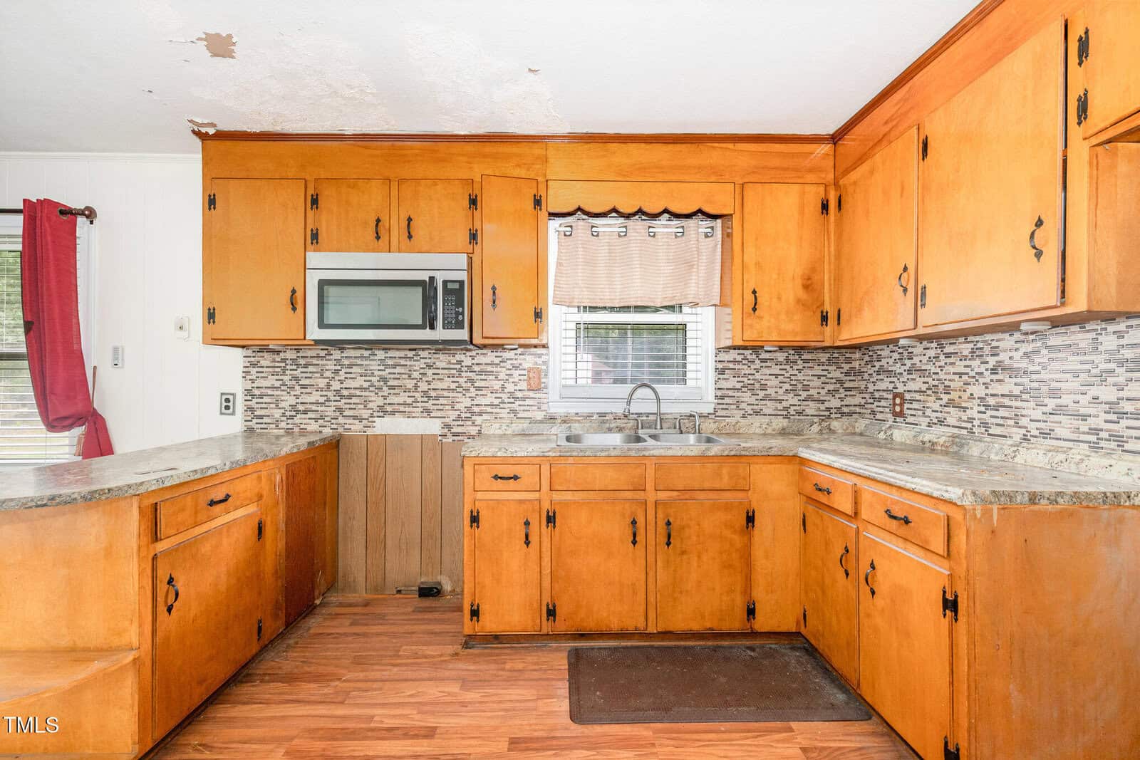 A kitchen with wood cabinets, a double sink under a window, a microwave built into the wall, stone-style countertops, mosaic tile backsplash, and wood flooring. The ceiling shows some peeling paint.