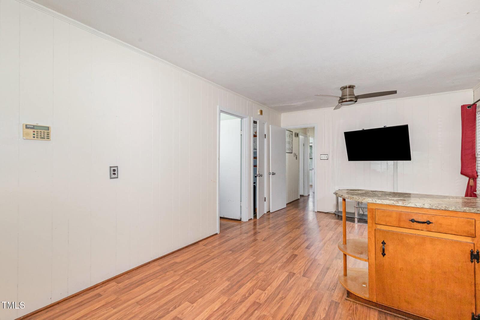 A room with light-colored walls, wood flooring, a ceiling fan, a wall-mounted TV, and a wooden cabinet with granite top. Several open doorways lead to other rooms.
