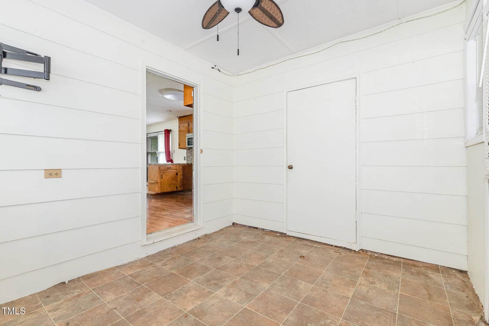 Small room with white paneled walls and tile floor, ceiling fan with leaf-shaped blades, and open doorway leading to a kitchen with wood cabinets and visible appliances. Closed white door on the right wall.