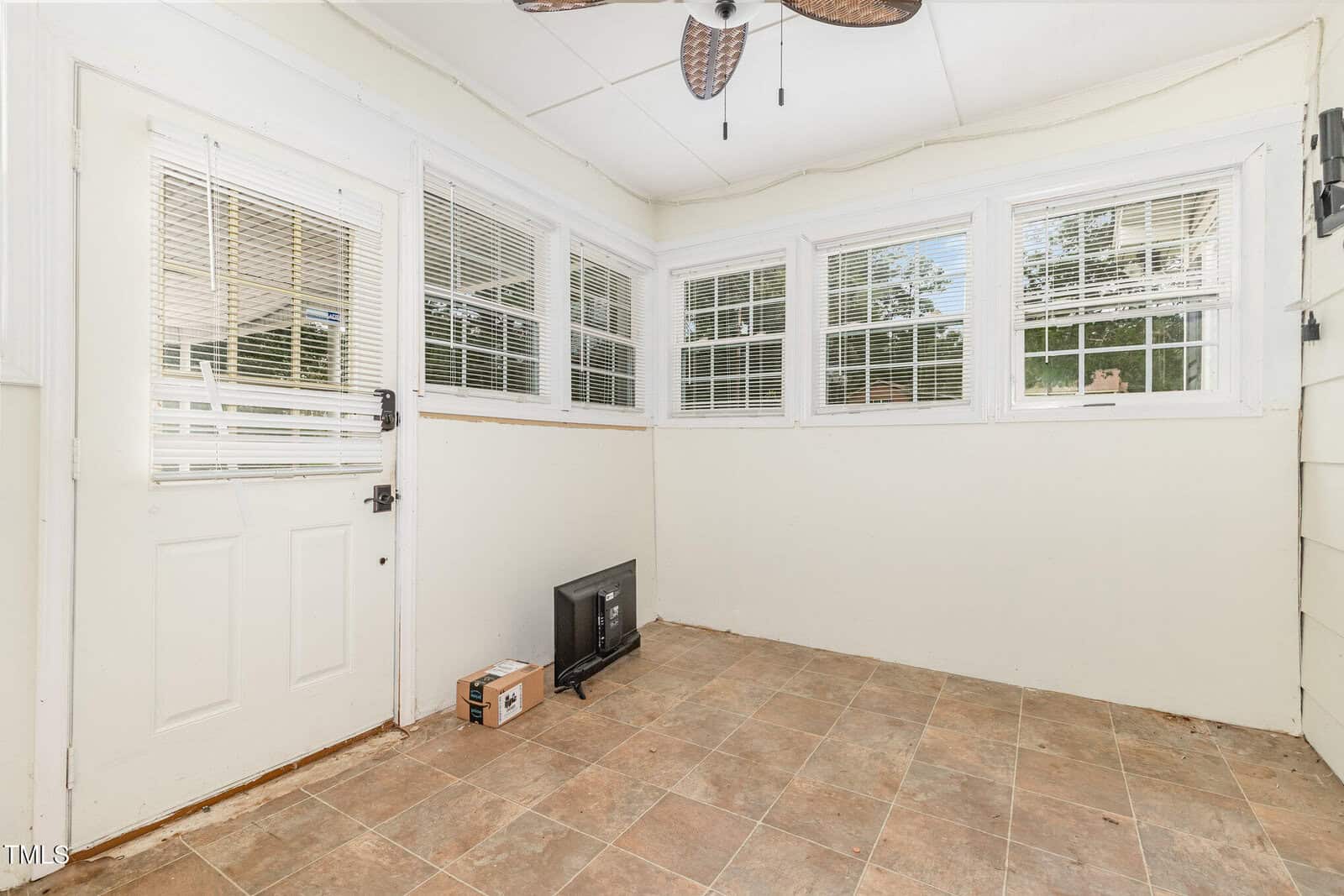A small, bright sunroom with tan tile flooring, cream-colored walls, a ceiling fan, multiple windows, a white door, and a TV and box on the floor near the wall.