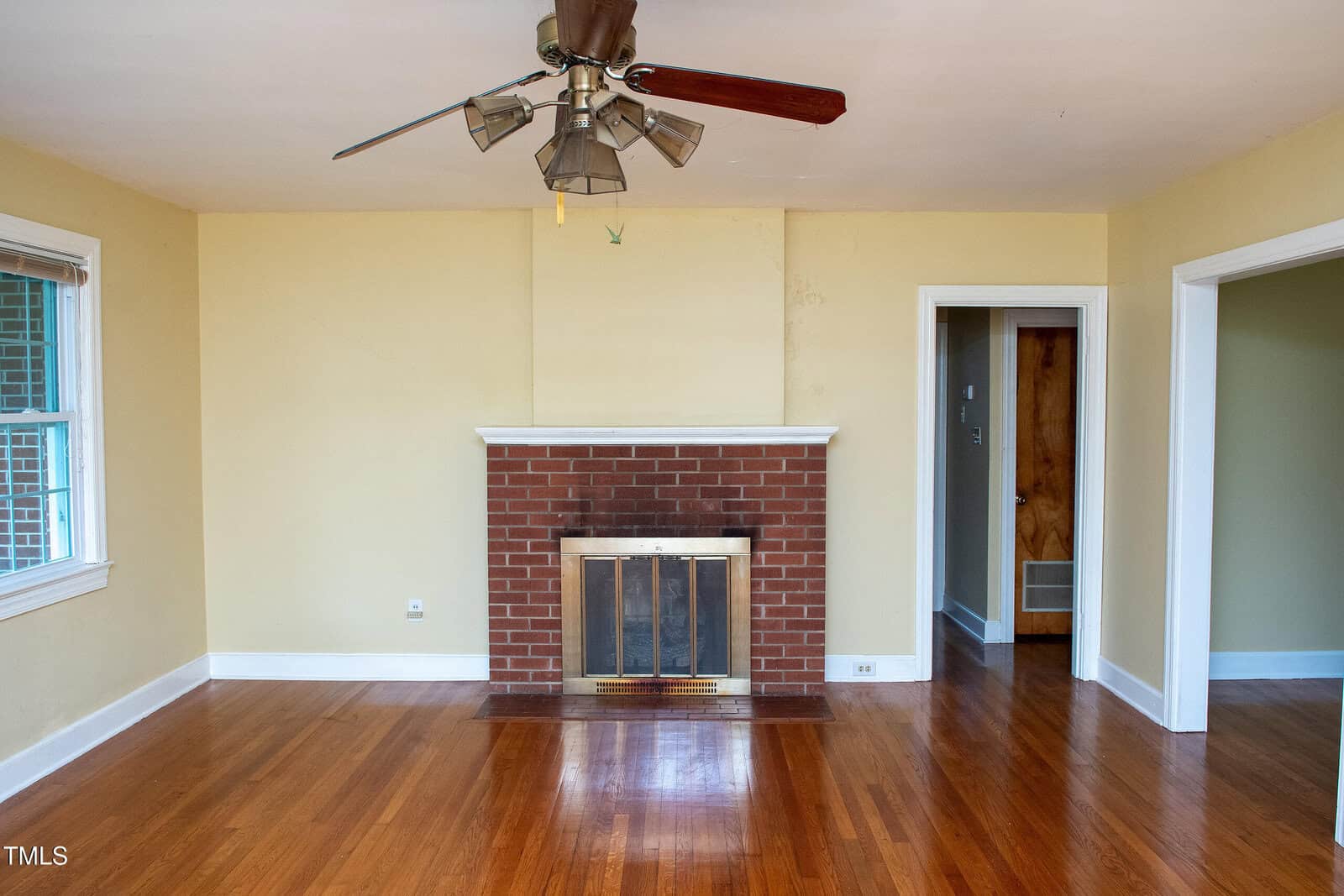 Empty living room with yellow walls, hardwood floors, brick fireplace with a brass screen, ceiling fan, two doorways, and a window on the left side.