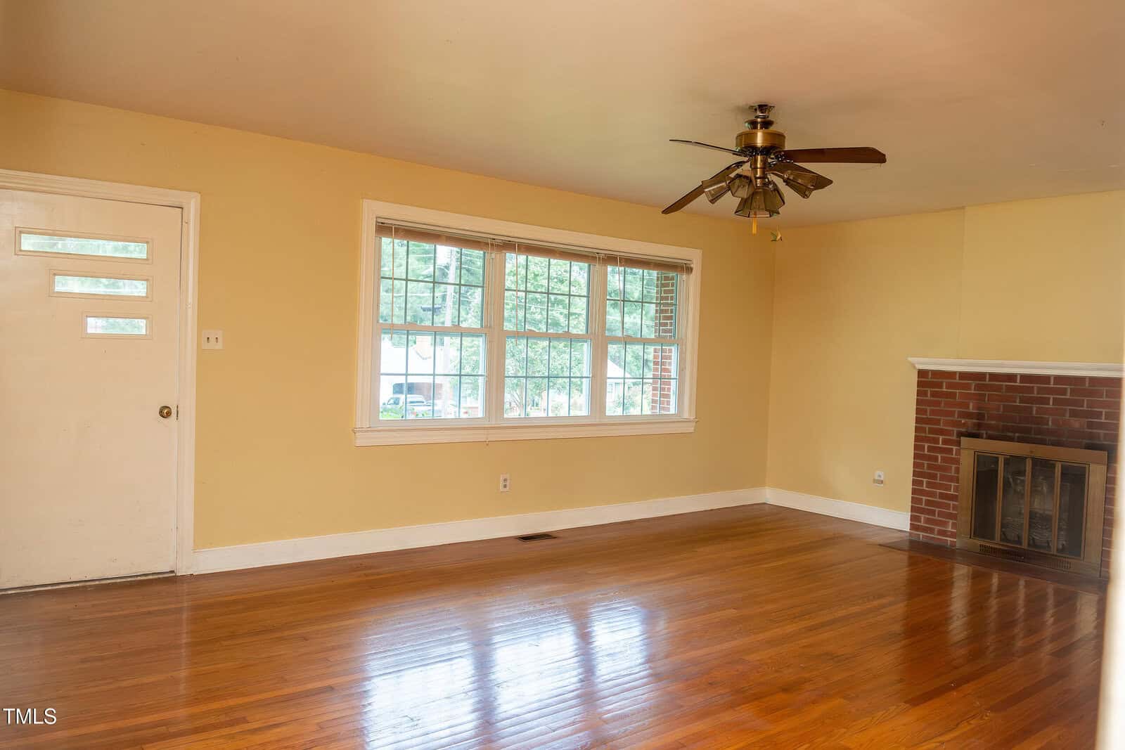 An empty living room with hardwood floors, a ceiling fan, large window, yellow walls, a white door, and a brick fireplace with a white mantel.