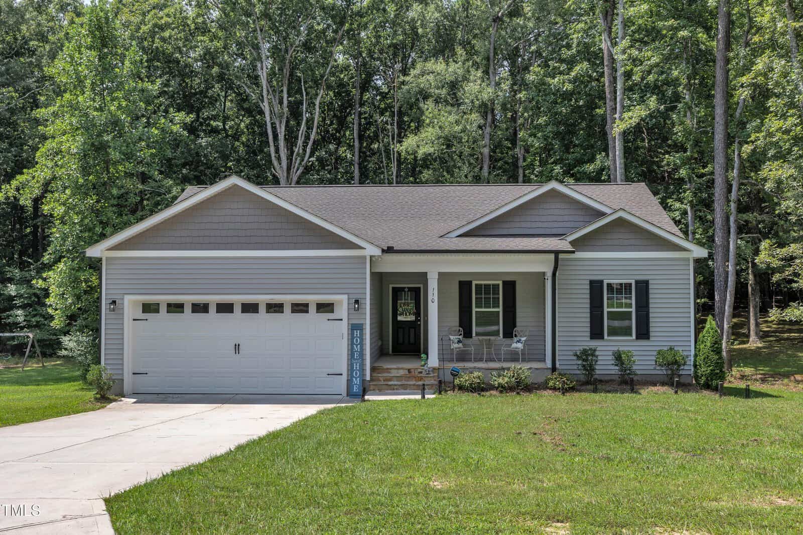 Single-story gray house with a two-car garage, front porch, and black shutters, set on a grassy lawn with small shrubs at 110 Allen Avenue in Franklinton. Tall trees and forest are visible in the background, while the driveway leads up to the garage.