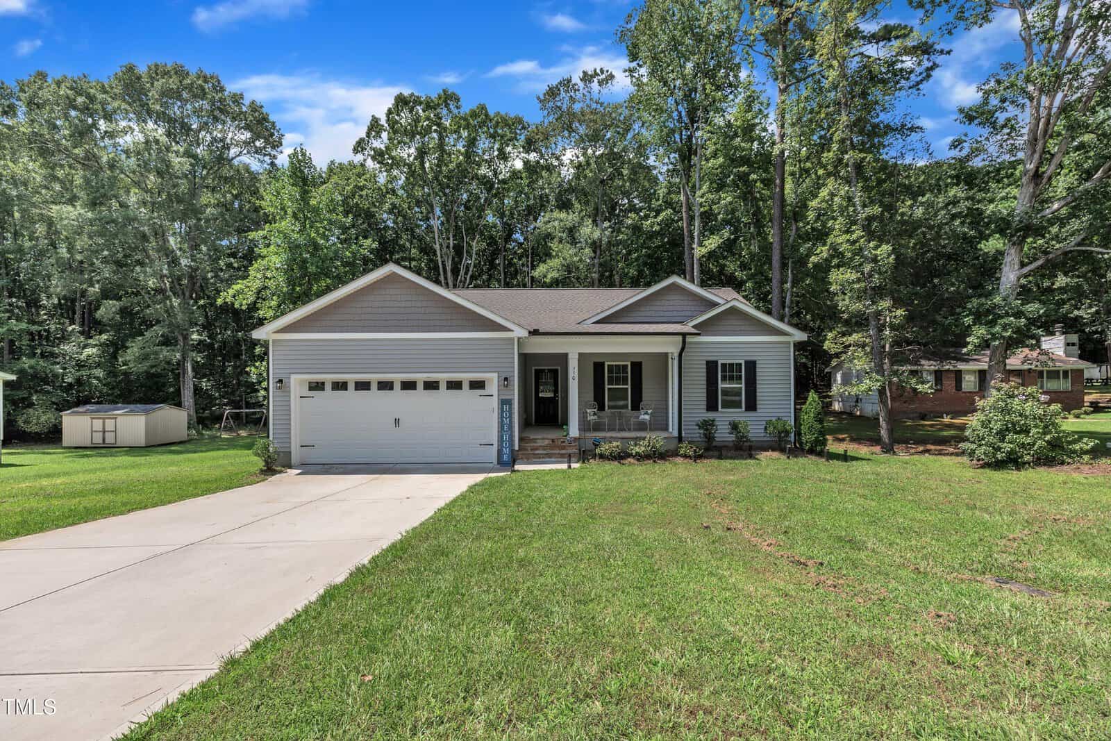 Single-story gray house with attached two-car garage, small front porch, and concrete driveway. Lawn and trees surround the home, with a shed visible to the left and another house partly visible in the background.