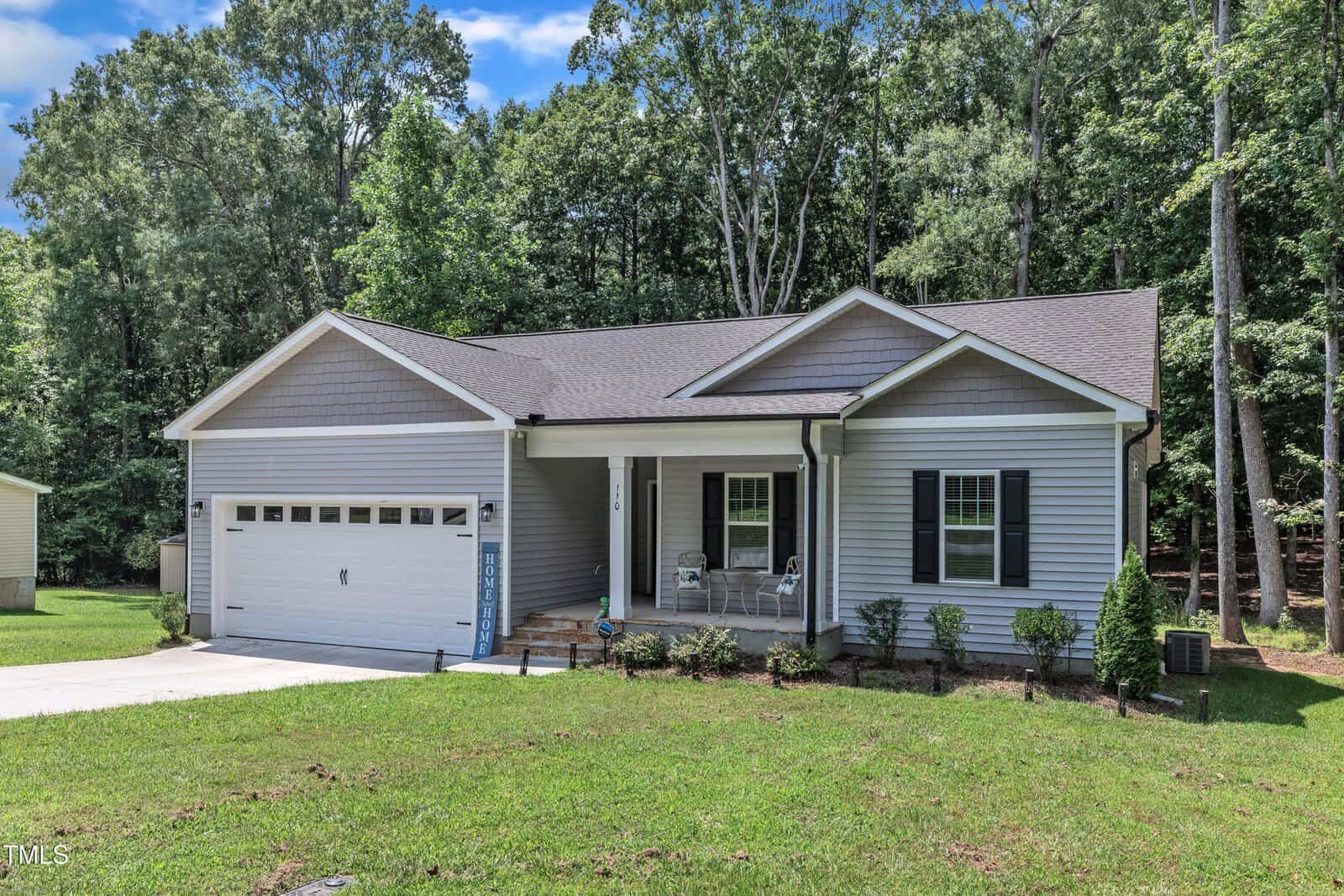 Single-story house with gray siding, white trim, and a covered front porch. There is a two-car garage on the left and a small front lawn with a few shrubs. The background features tall trees and a blue sky.
