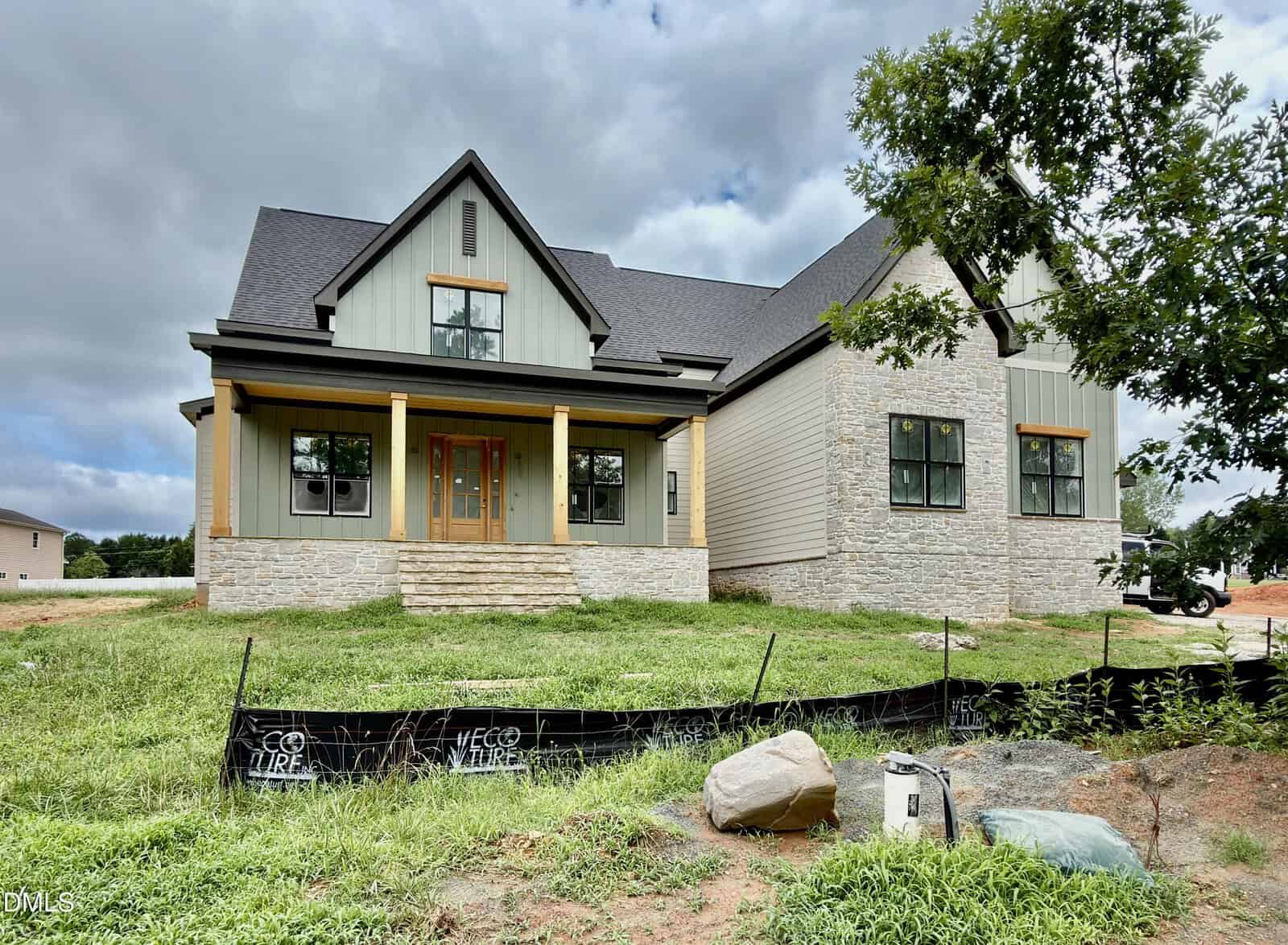 A newly constructed modern farmhouse at 1121 Dovefield Lane in Youngsville features gray siding, stone accents, wooden pillars, and a covered front porch on a grassy lot. Construction materials and erosion fencing are visible in the yard under a cloudy sky.