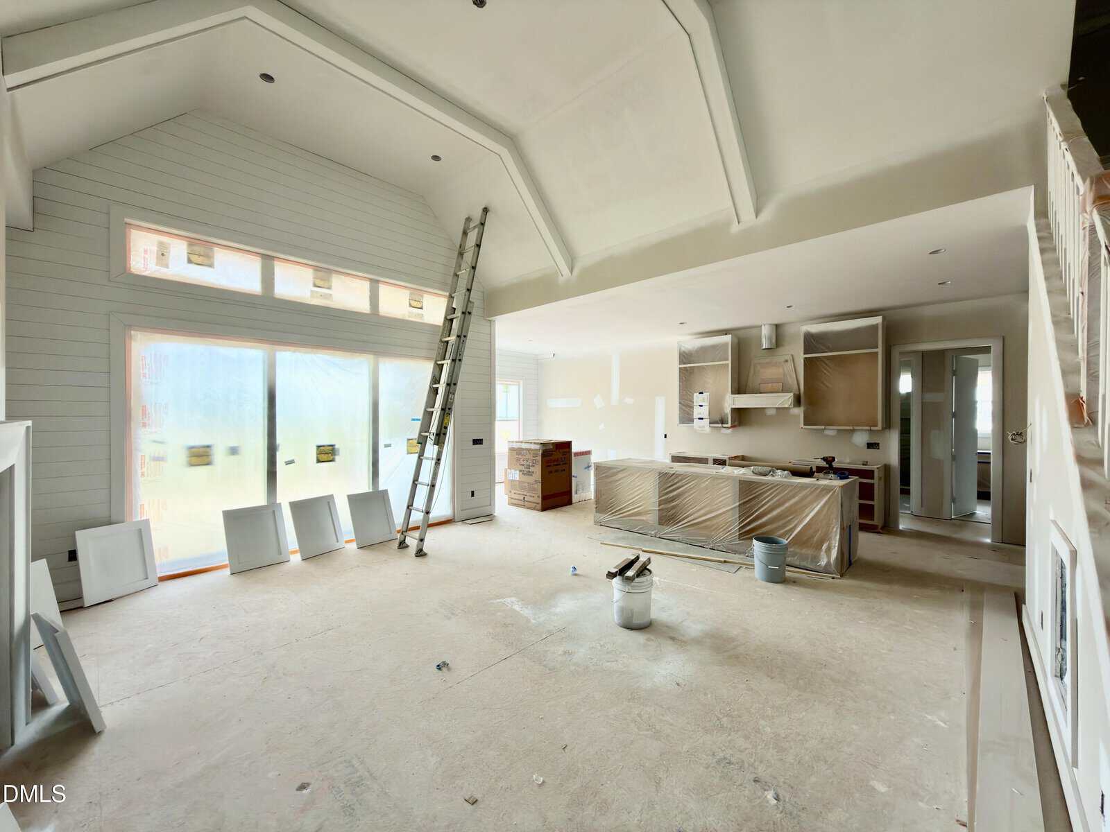 A modern kitchen and living area under construction, with a high vaulted ceiling, ladder, unfinished floors, covered cabinets, tools, and materials scattered around, and large windows letting in natural light.