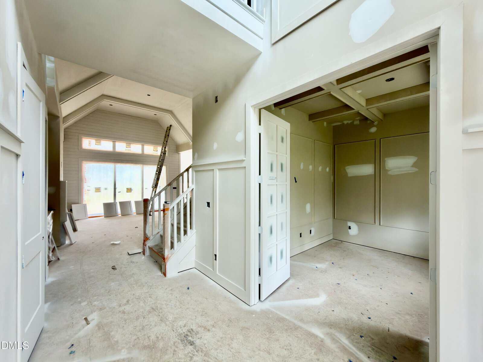 Interior of a house under construction, featuring unfinished walls with drywall patches, a staircase with protective covering, scattered construction materials, and a room with double doors partially open. Natural light enters from large windows.