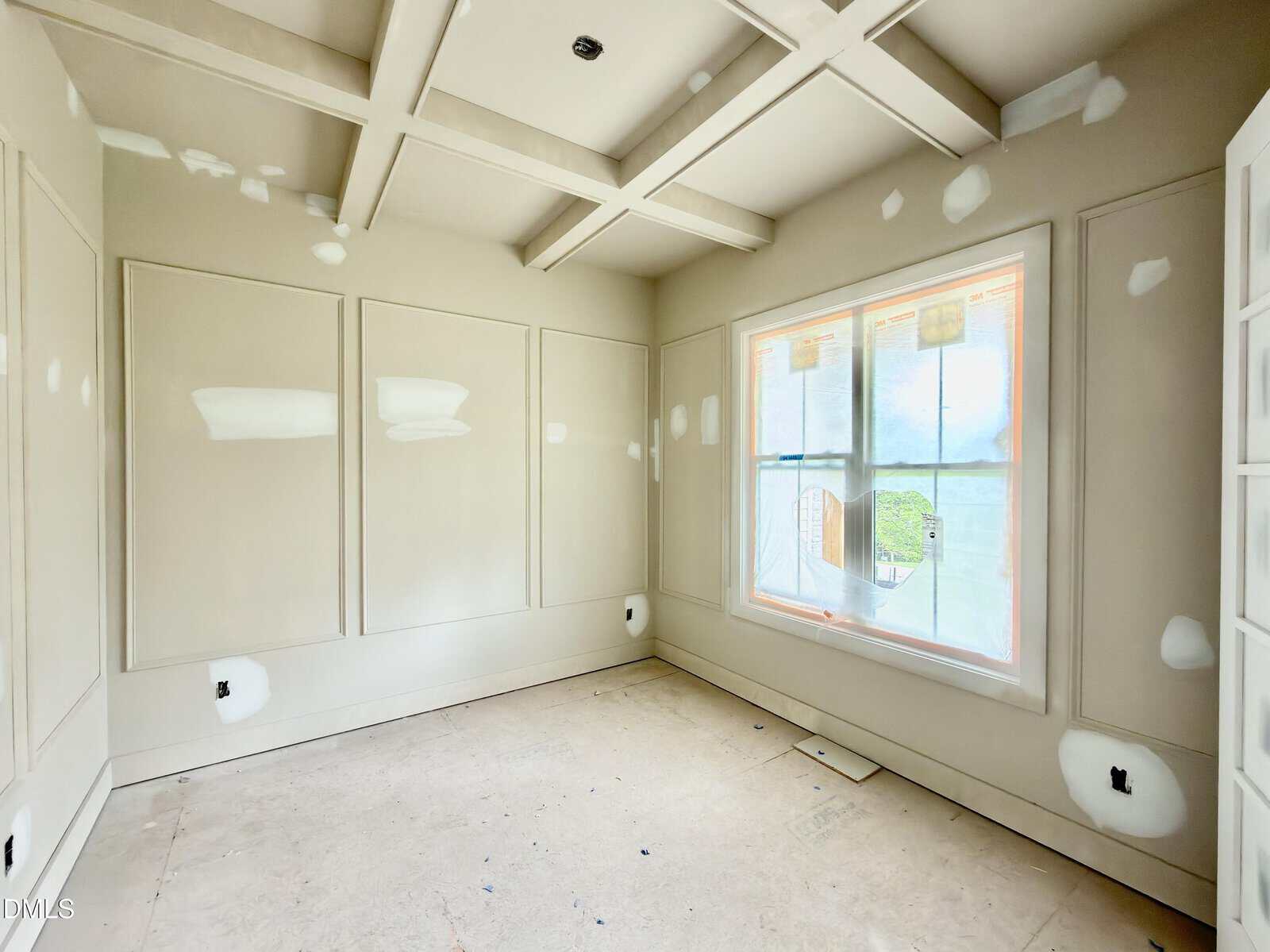 A bright, unfinished room with beige walls, coffered ceiling, large window, and white patches of spackle on the walls and trim, indicating ongoing construction or painting work.
