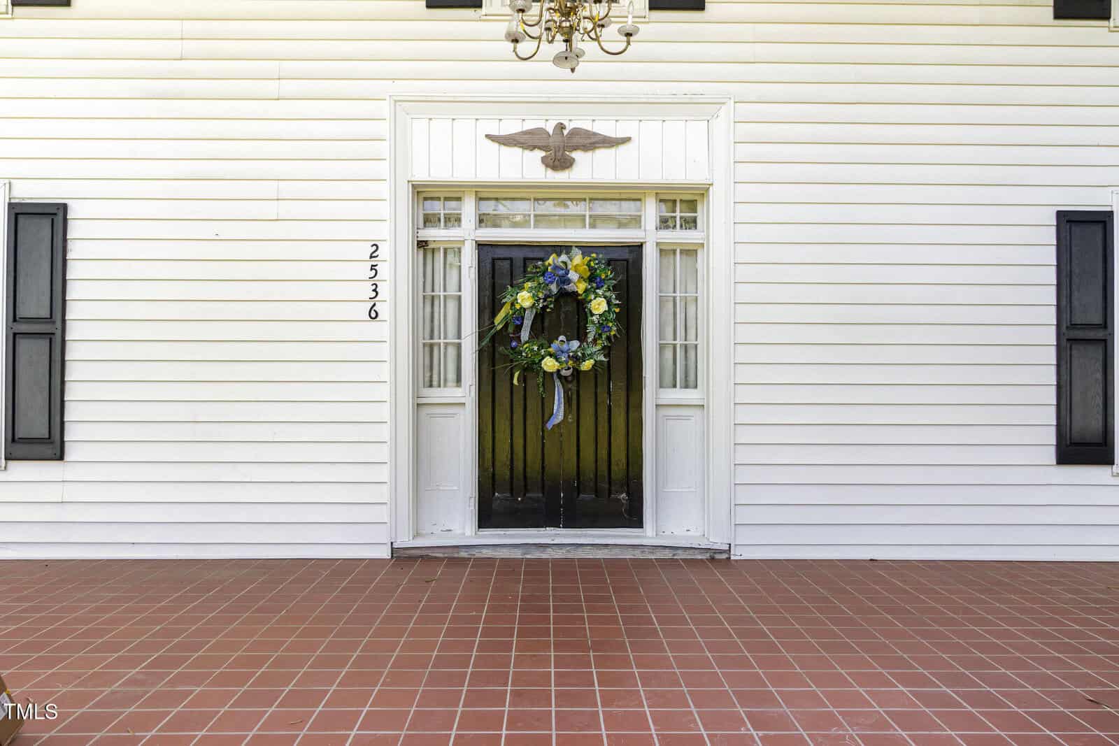 A white front door with glass panels, flanked by sidelights, features a large decorative wreath. The house has white siding, black shutters, and a red tile porch floor. House number 2536 is mounted vertically by the door.