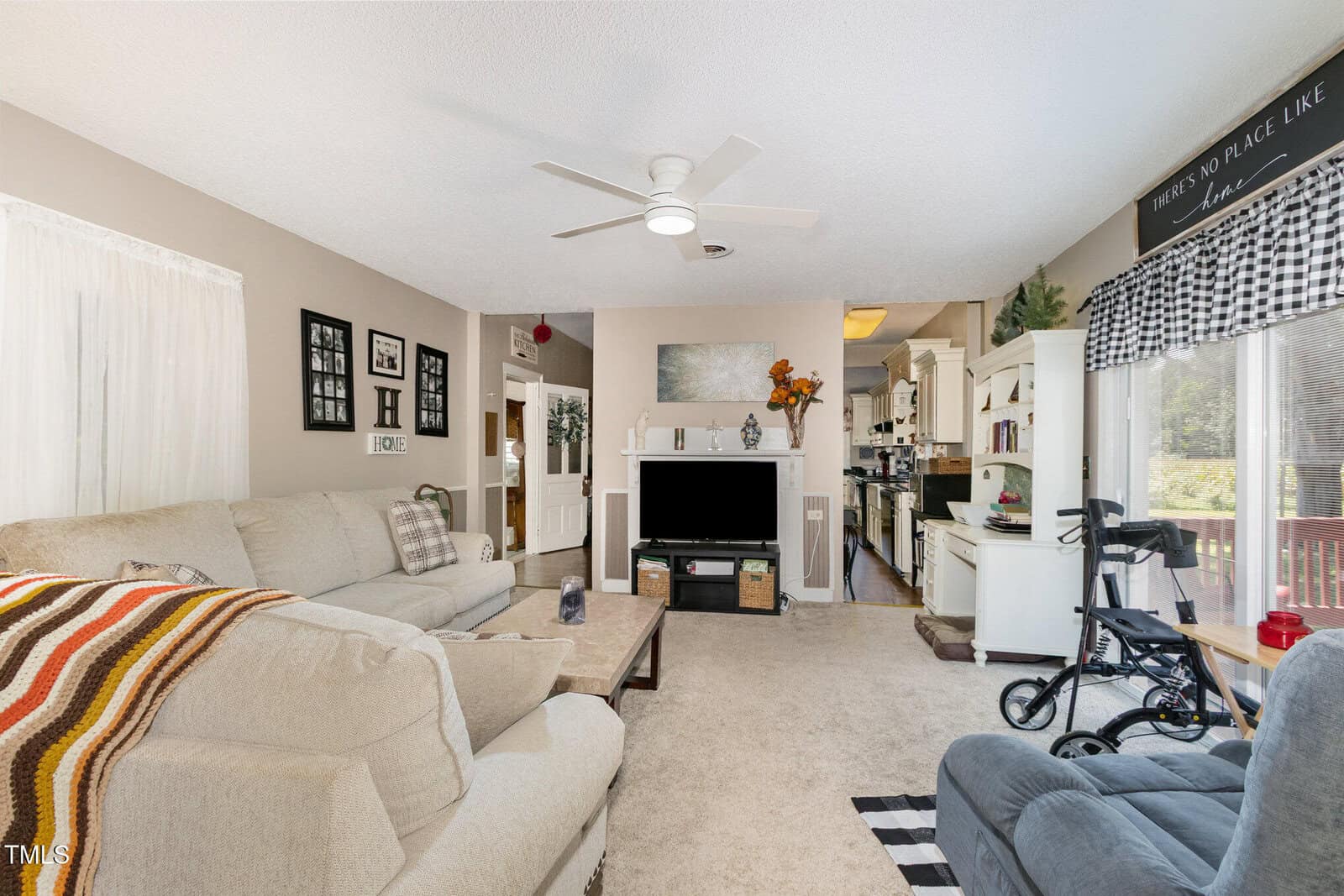 A living room with beige carpet and walls, a sectional sofa with a striped blanket, a coffee table, a fireplace, a ceiling fan, and a view into a kitchen; a walker is near a glass door to a deck.