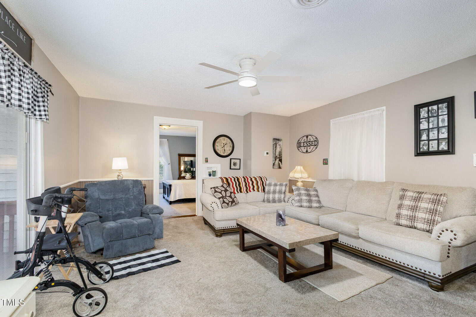 A living room with beige walls and carpet, a light-colored sectional sofa, a recliner, a walker, a wooden coffee table, framed photos on the wall, and a window with checkered curtains. A doorway leads to another room.