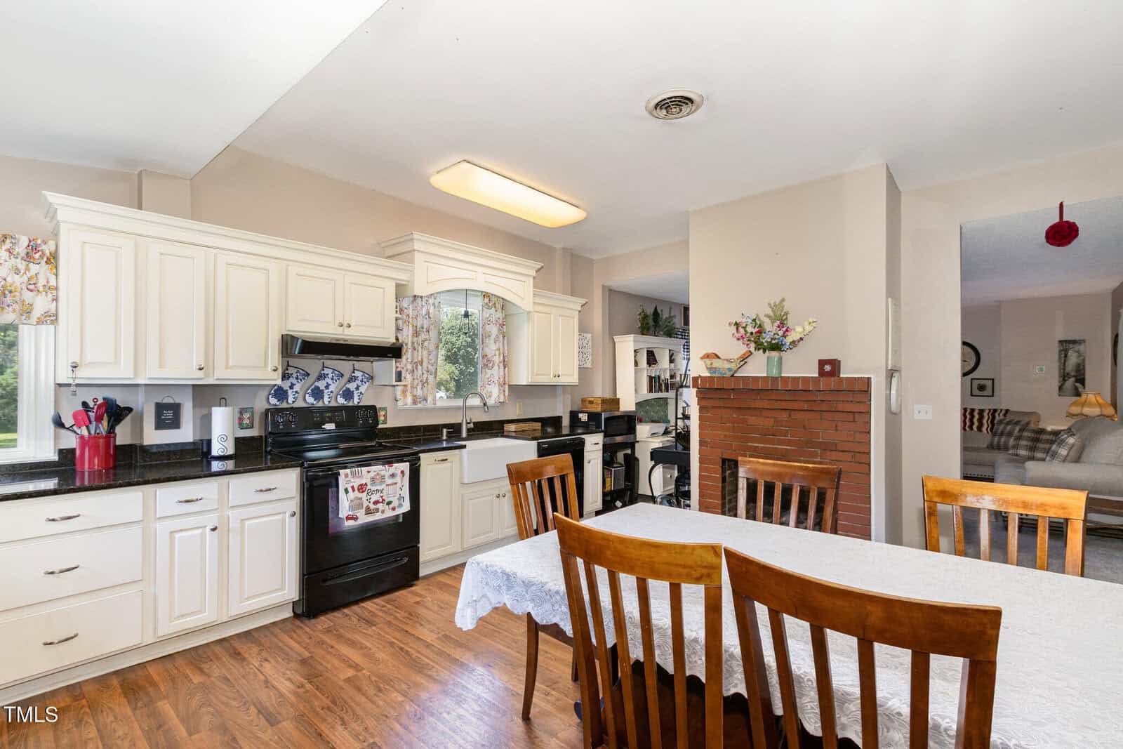 Bright kitchen with white cabinets, black appliances, and a dining table with a white tablecloth. A partial brick divider separates the kitchen from a living room area with a sofa and visible decor.