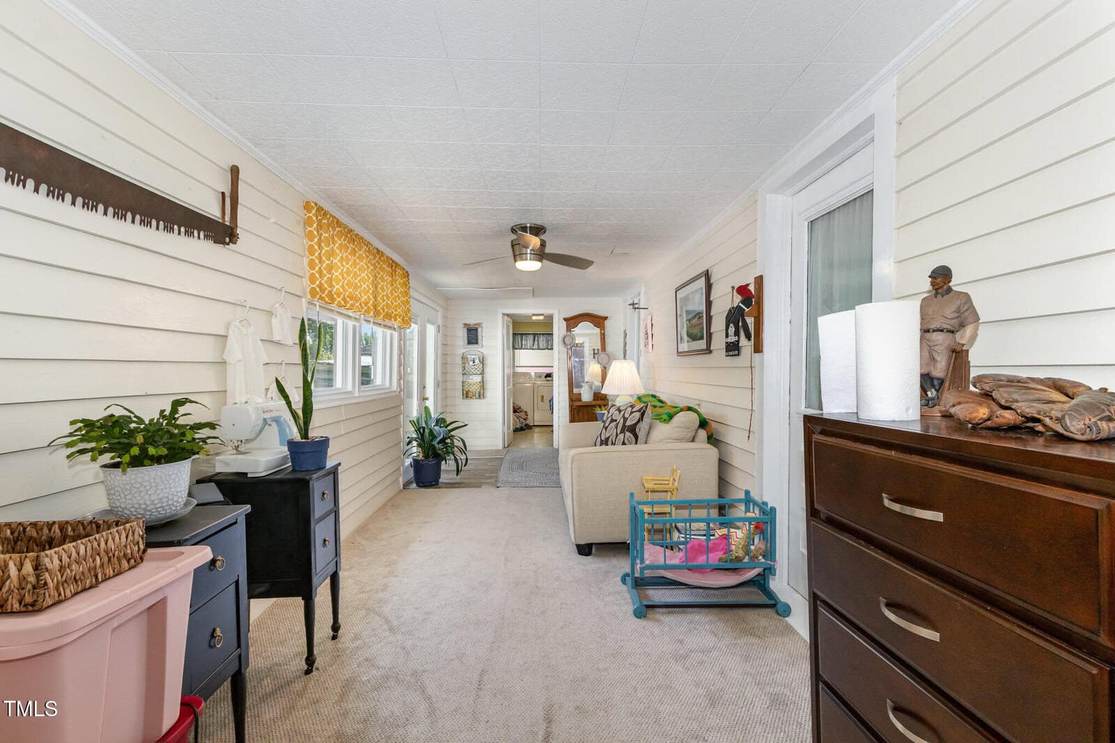Bright sunroom with light walls, carpeted floor, couch, side tables, plants, ceiling fan, yellow curtains, dresser, and various decorative items. Large windows line one side, allowing natural light to fill the space.