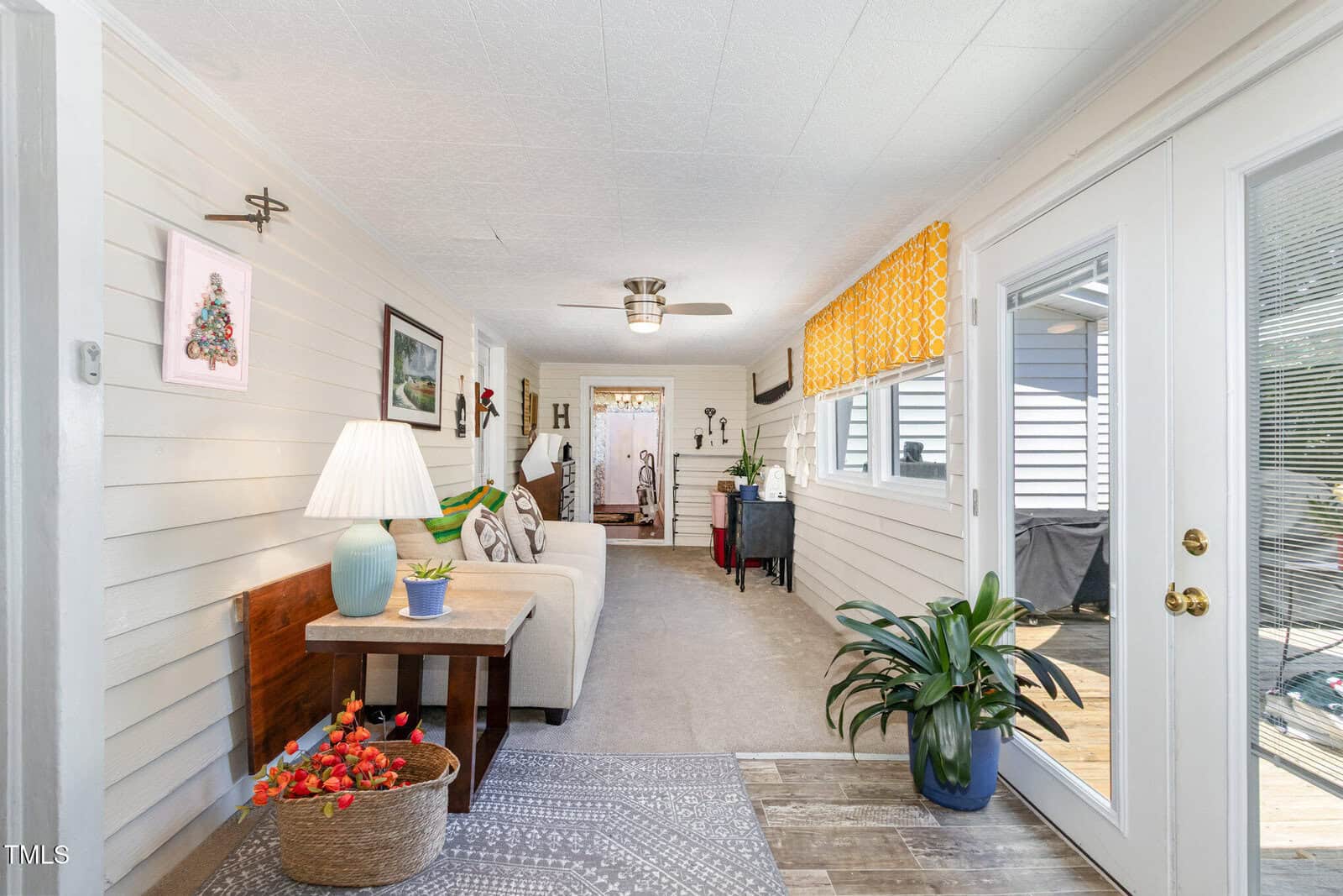 Bright sunroom with white paneled walls, beige sofa, lamp, small plant, and a glass door leading outside. A patterned rug and large potted plant complete the cozy, inviting space.