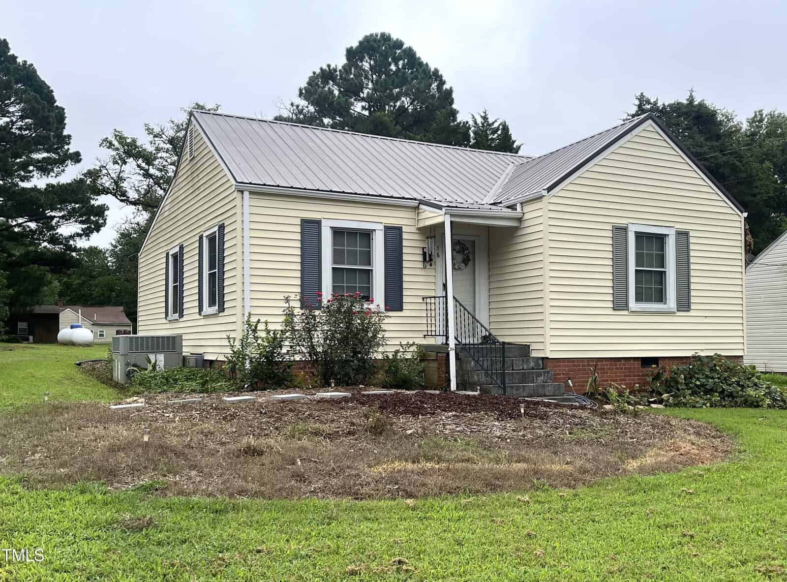 A small, single-story beige house with gray shutters and a metal roof sits on a grassy yard. There are bushes near the front steps and a propane tank visible in the background. Trees surround the property.