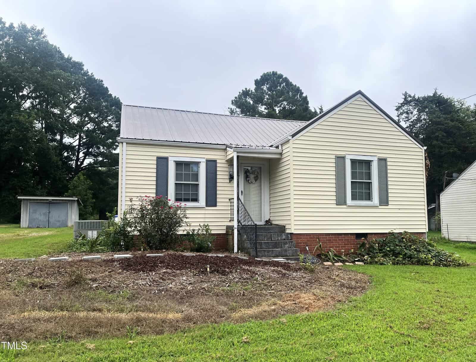Small, single-story beige house with gray shutters and a metal roof. The front door is accessed by steps with railings, and there is a shed in the backyard. The yard has grass, bushes, and a mulched area. Trees surround the property.