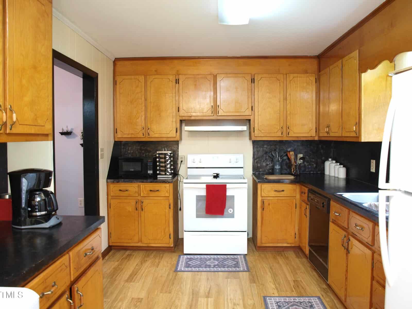 A kitchen with wooden cabinets, a white stove and oven, white refrigerator, black countertop appliances, a dishwasher, and two small rugs on a wood floor. A red towel hangs on the oven handle.