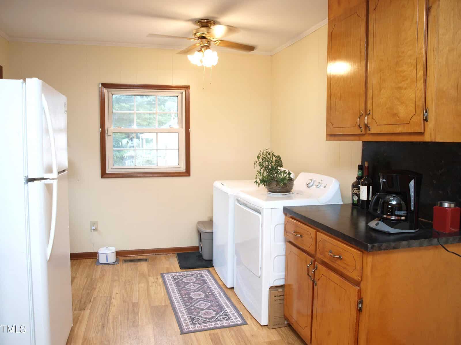 A kitchen with wood cabinets, a black countertop, a coffee maker, and a washer and dryer. A potted plant sits on the dryer. There is a window, ceiling fan, and a rug on the wood floor.