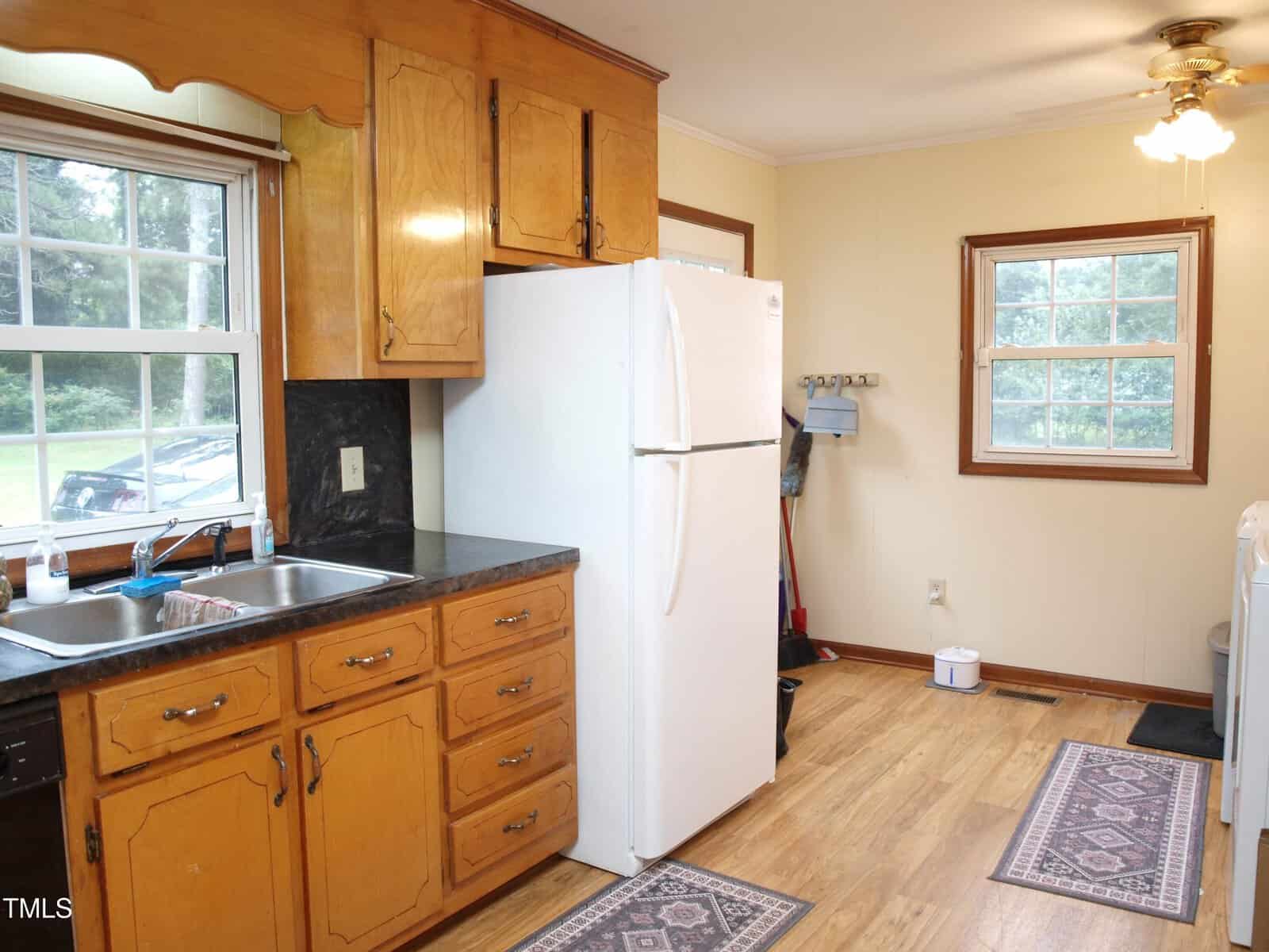A kitchen with wooden cabinets, a white refrigerator, black countertop, stainless steel sink under a window, and wood flooring. Two rugs are on the floor and a window lets in natural light.