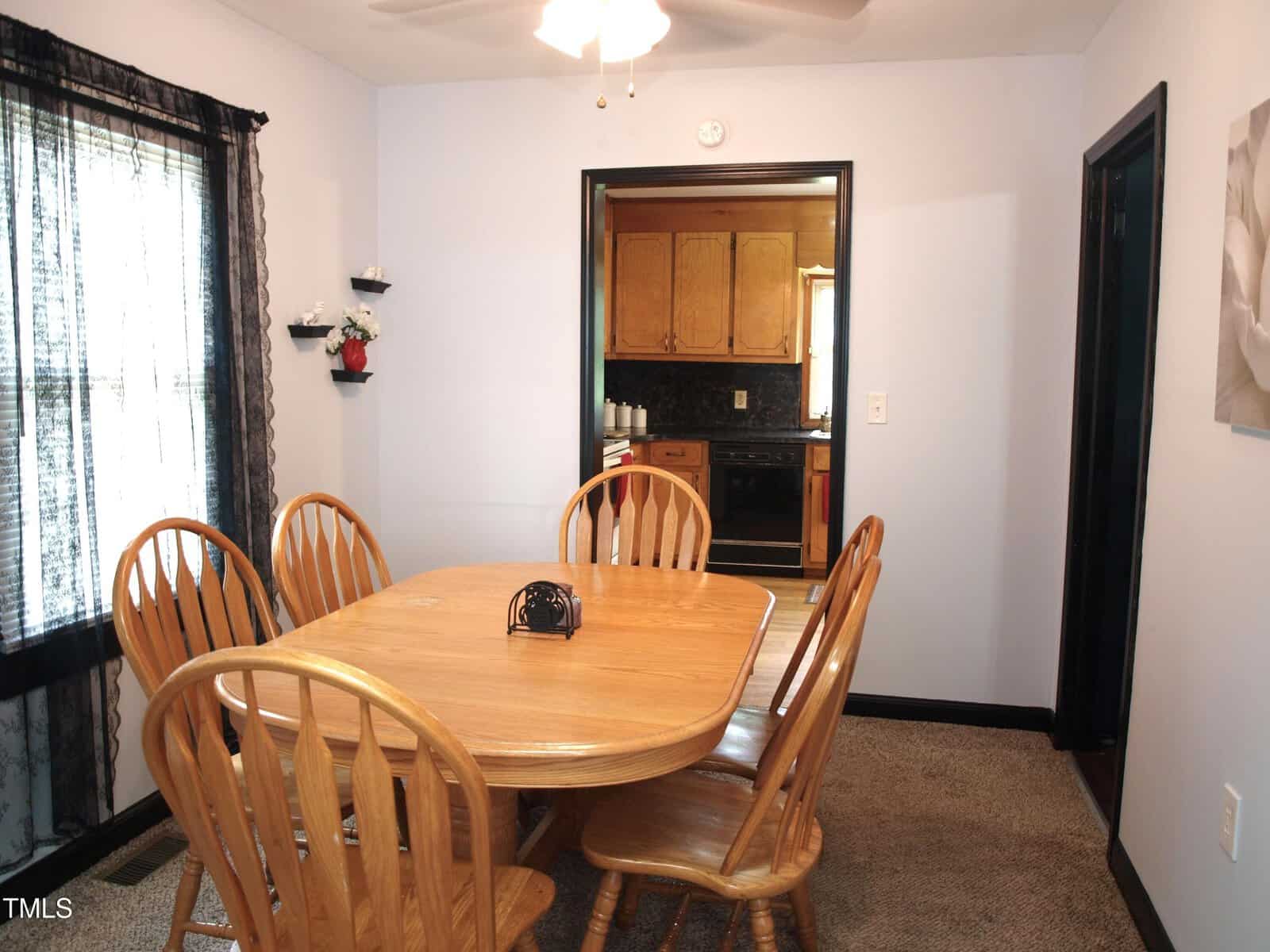 A dining room with a rectangular wooden table and six matching chairs. A window with black lace curtains is on the left, and a doorway leads to a kitchen with wooden cabinets and a black countertop.
