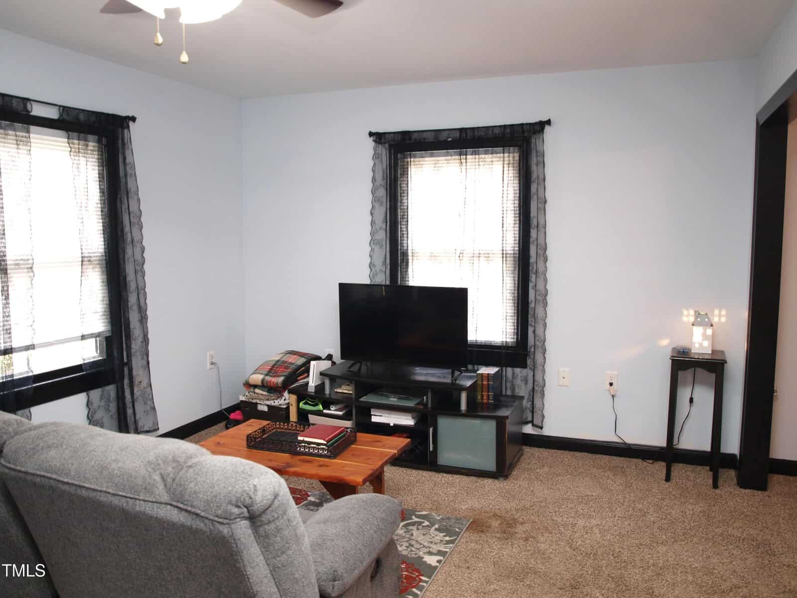 A living room with light blue walls, two windows with black curtains, a TV on a stand, a gray armchair, a wooden coffee table with items on it, and a small lamp on a side table. Brown carpet covers the floor.