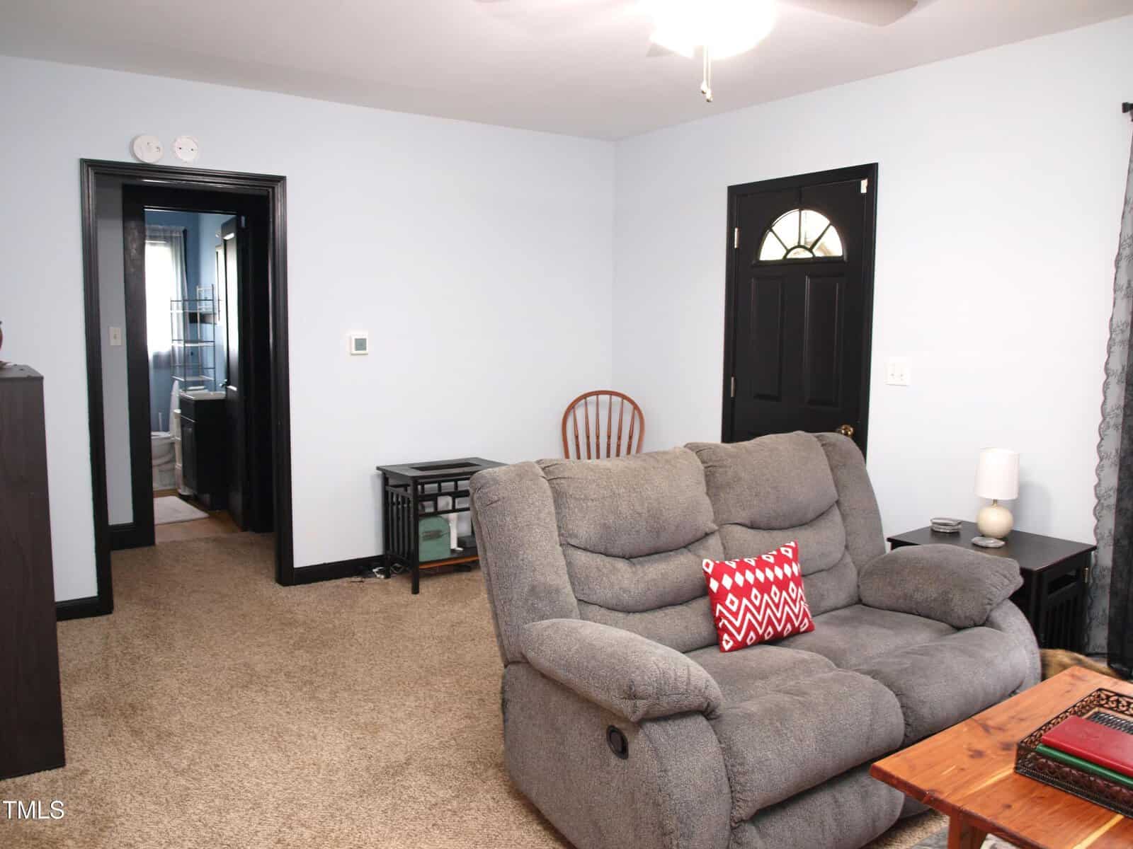 A living room with beige carpet, a gray reclining loveseat, a small wooden chair, a black side table, and a black door. There is a lamp and decor items on side tables, and a doorway leads to another room.