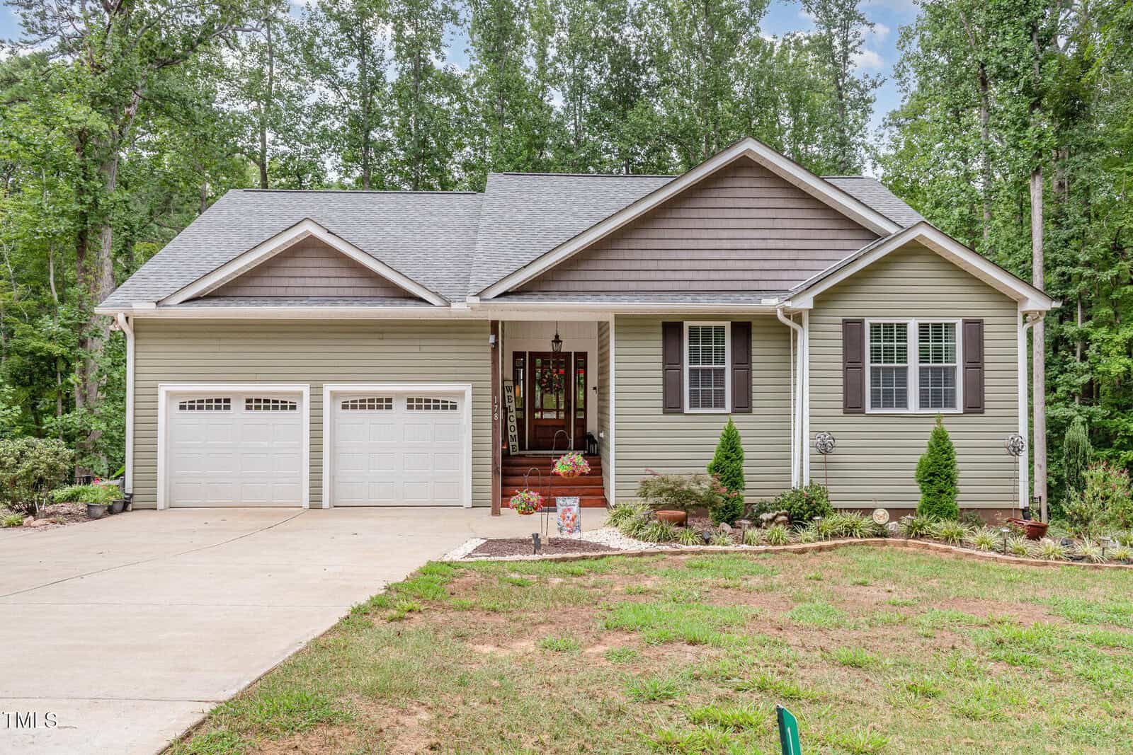 Single-story house with gray siding, double garage, small porch with potted plants, front lawn, landscaped garden beds, and surrounded by tall trees in the background—nestled on 178 Horseman Drive in the charming community of Louisburg.