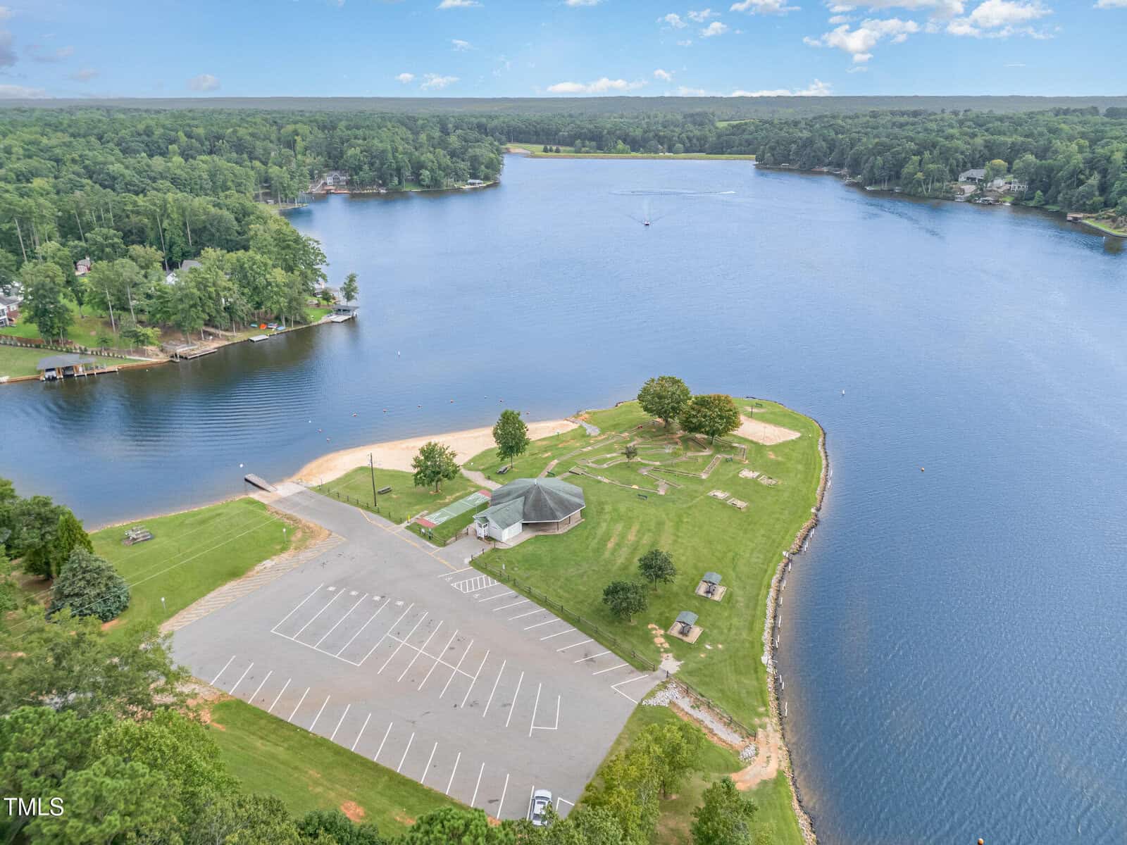 Aerial view of a lakeside park with a sandy beach, grassy areas, picnic tables, a pavilion, playground, and an adjacent empty parking lot surrounded by trees. The lake extends into the distance under a partly cloudy sky.