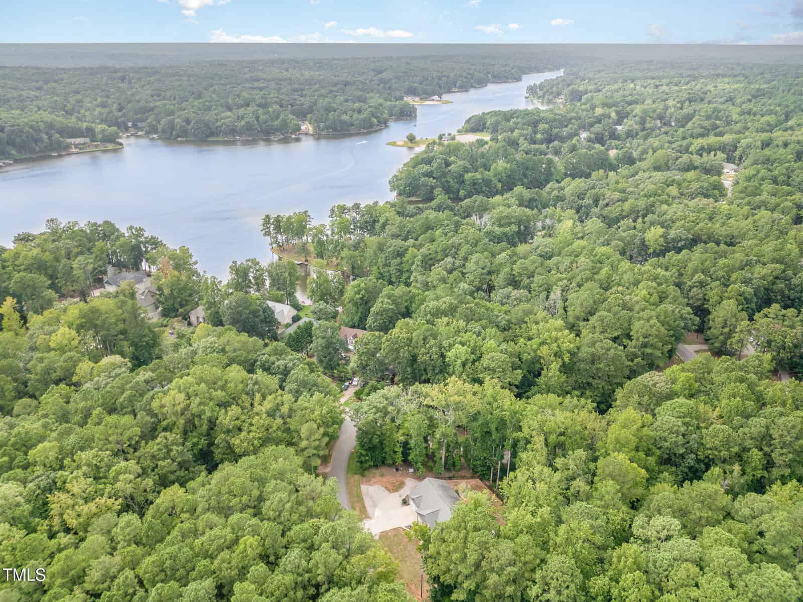 Aerial view of a house surrounded by dense trees near a large lake, with several other homes and wooded areas visible along the shoreline and in the background.