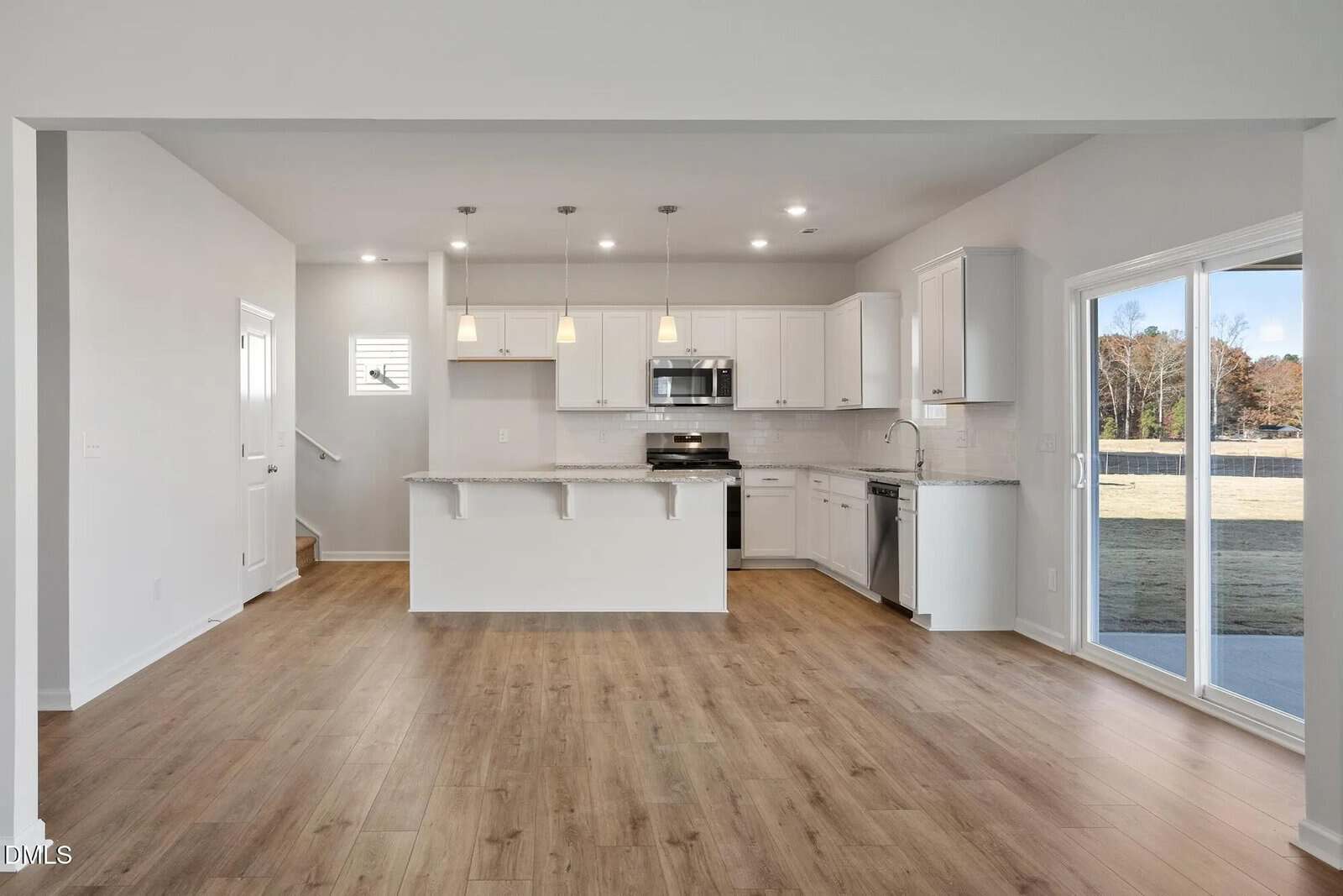 Modern kitchen with white cabinets, stainless steel appliances, pendant lights, and a kitchen island. Open layout with wood-style flooring and large sliding glass doors leading outside. Walls are light gray, and space is unfurnished.
