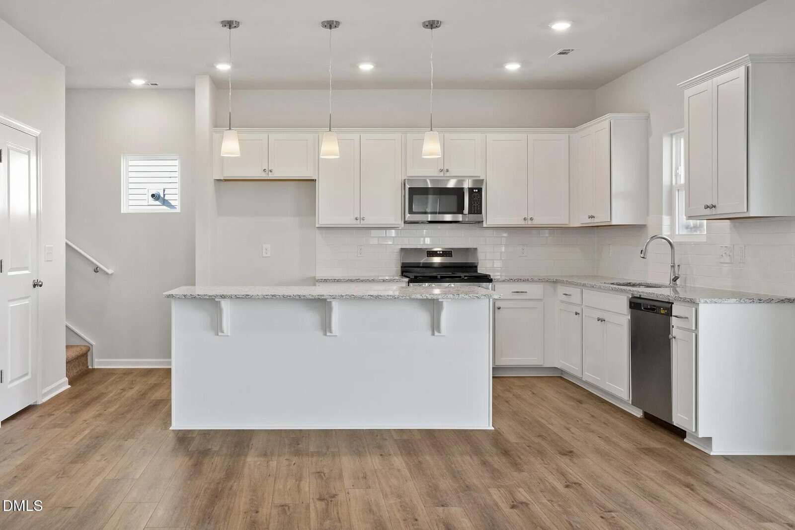 Bright, modern kitchen with white cabinets, granite countertops, stainless steel appliances, pendant lights over a large island, and wood-style flooring. Stairs are visible to the left, and there are two small windows.