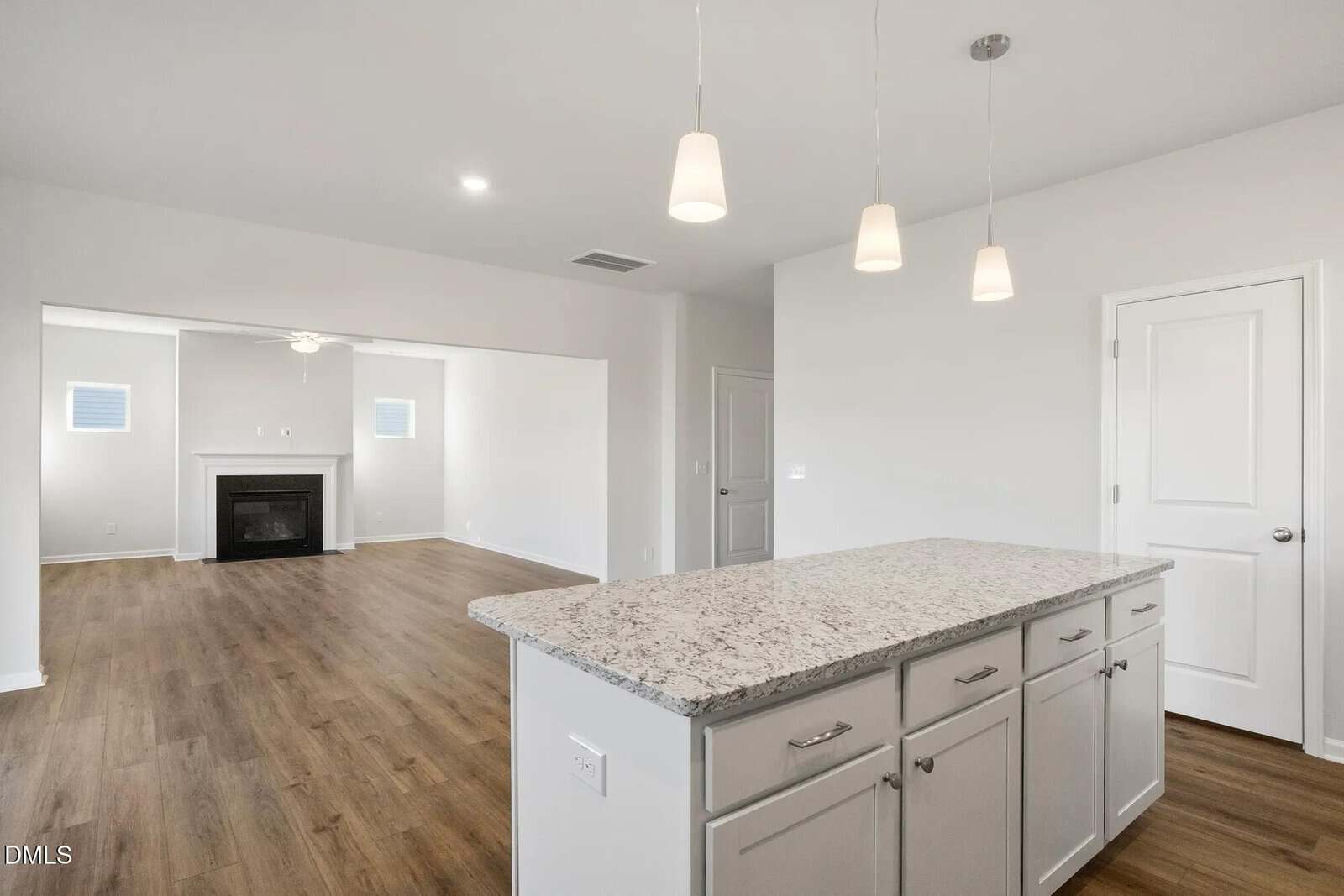 A modern kitchen with a granite island, white cabinets, and pendant lights opens to a living area with wood flooring, a white fireplace, and windows on either side. Two closed doors are visible. The walls are painted light gray.