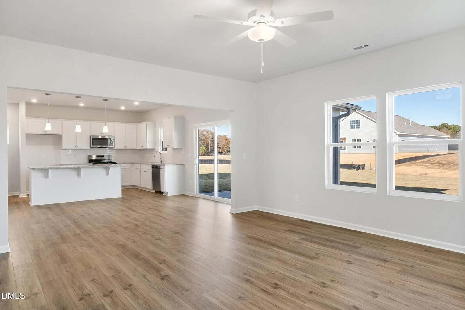 Bright, empty open-concept room with light wood floors, white walls, ceiling fan, and large windows. Kitchen in back has white cabinets, stainless steel appliances, pendant lights, and a sliding glass door to a backyard.