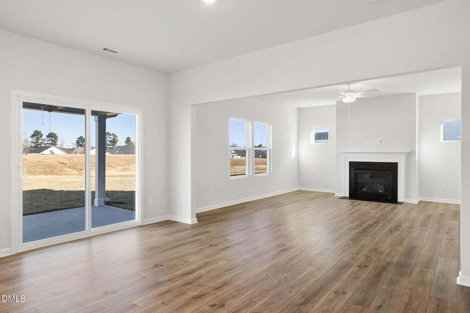 Bright, unfurnished living room with wood floors, a white ceiling fan, fireplace, multiple windows, and a sliding glass door leading to a backyard with grass and trees visible outside.