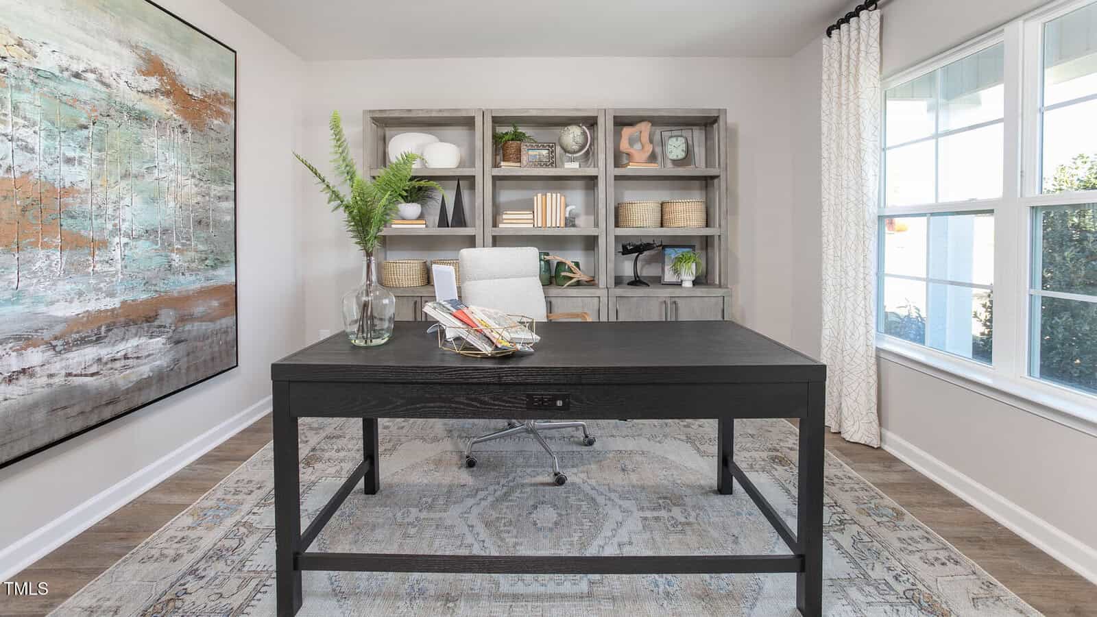 A modern home office with a dark wood desk, a gray office chair, a bookshelf with decor and baskets, a large abstract painting on the wall, a vase with ferns on the desk, and a window with patterned curtains.