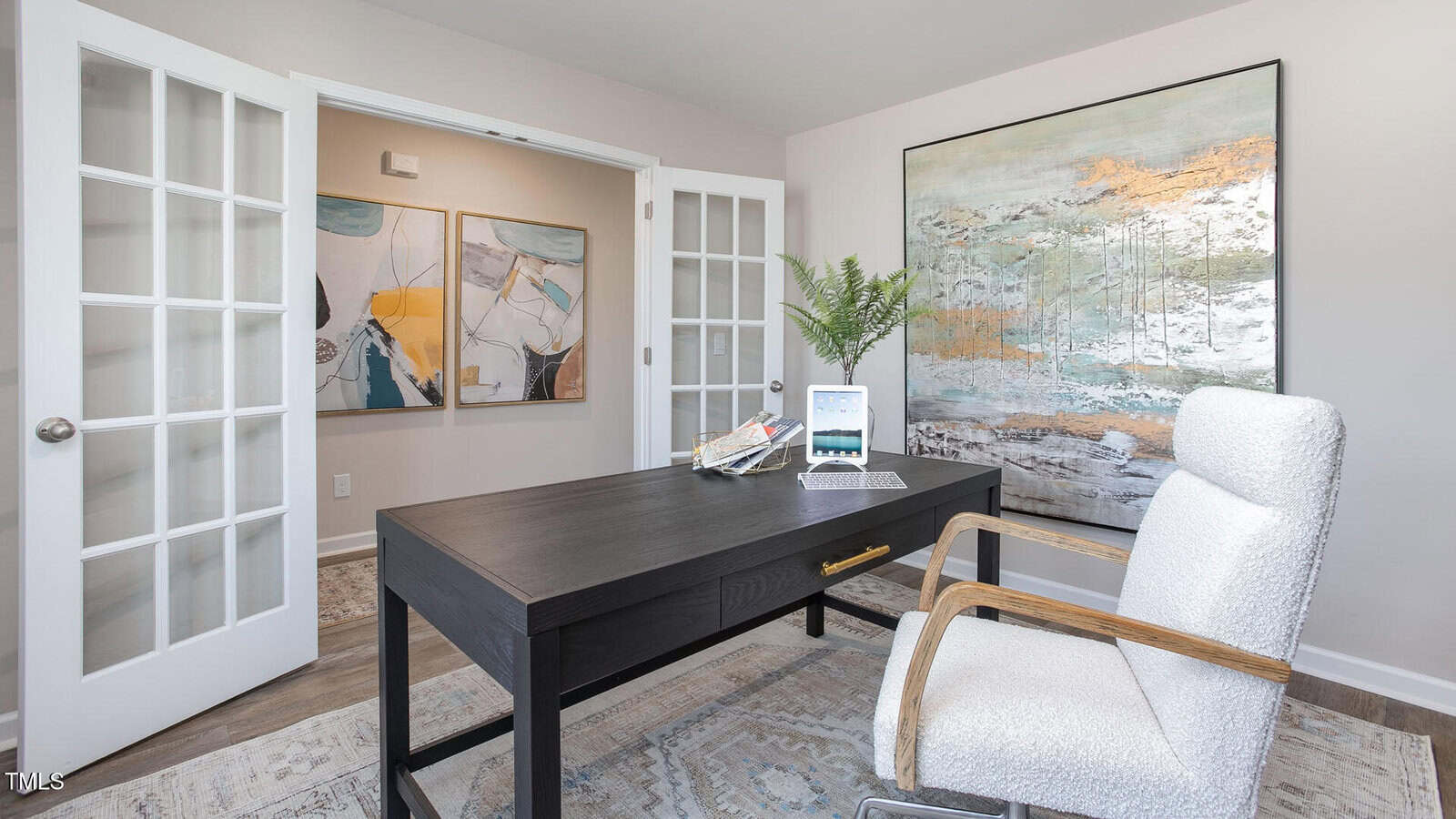 A modern home office features a black desk with a laptop and books, a white upholstered chair, wall art, and double French doors leading to an adjacent hallway with additional abstract artwork.